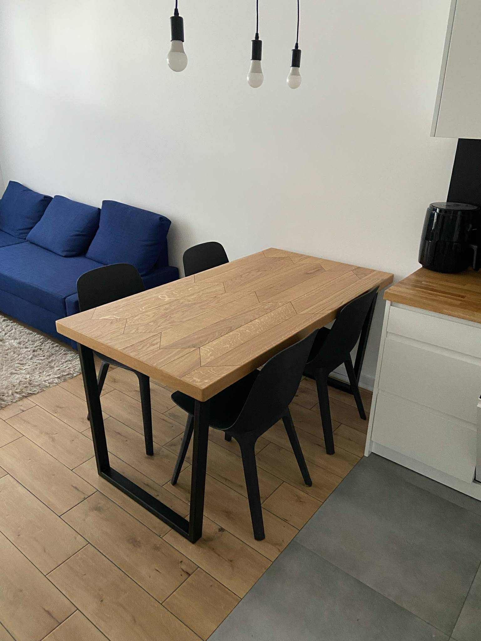 Drewniany stół z czarnymi nogami i krzesłami w nowoczesnym wnętrzu. Parkiet w ciepłym odcieniu, niebieska sofa i minimalistyczne lampy wiszące.