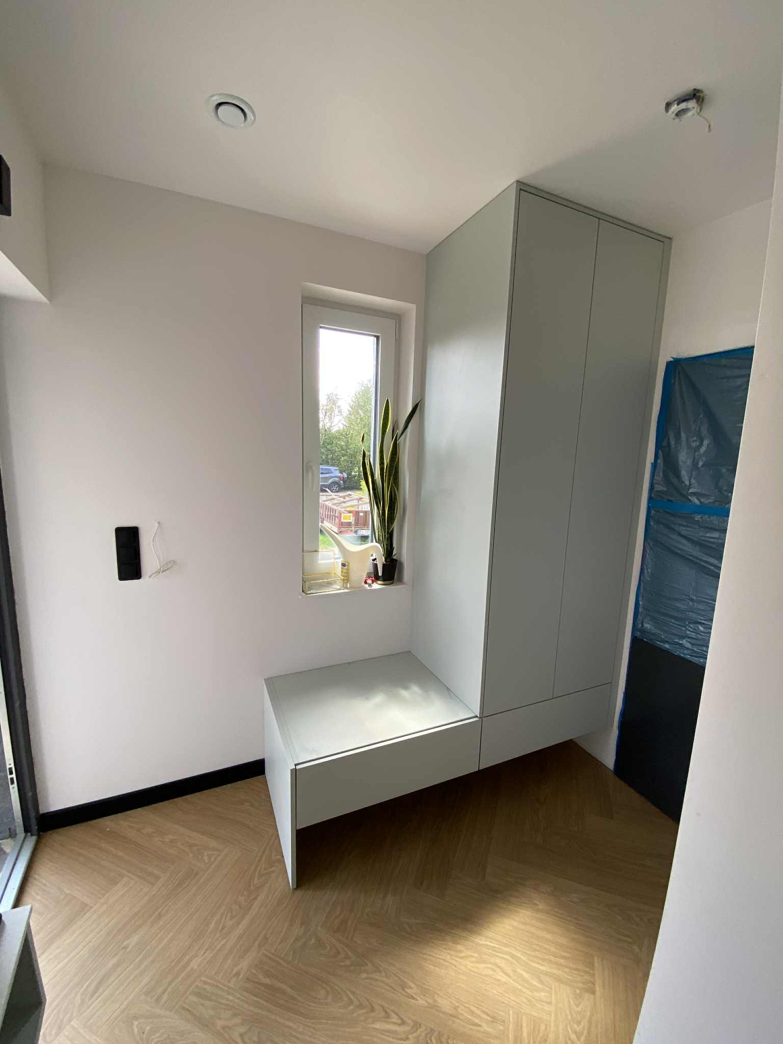 Jasna szafa z siedziskiem wnękowym przy oknie, z rośliną. Parkiet ułożony w jodełkę. Drzwi zabezpieczone folią malarską. Minimalistyczny design wnętrza.
