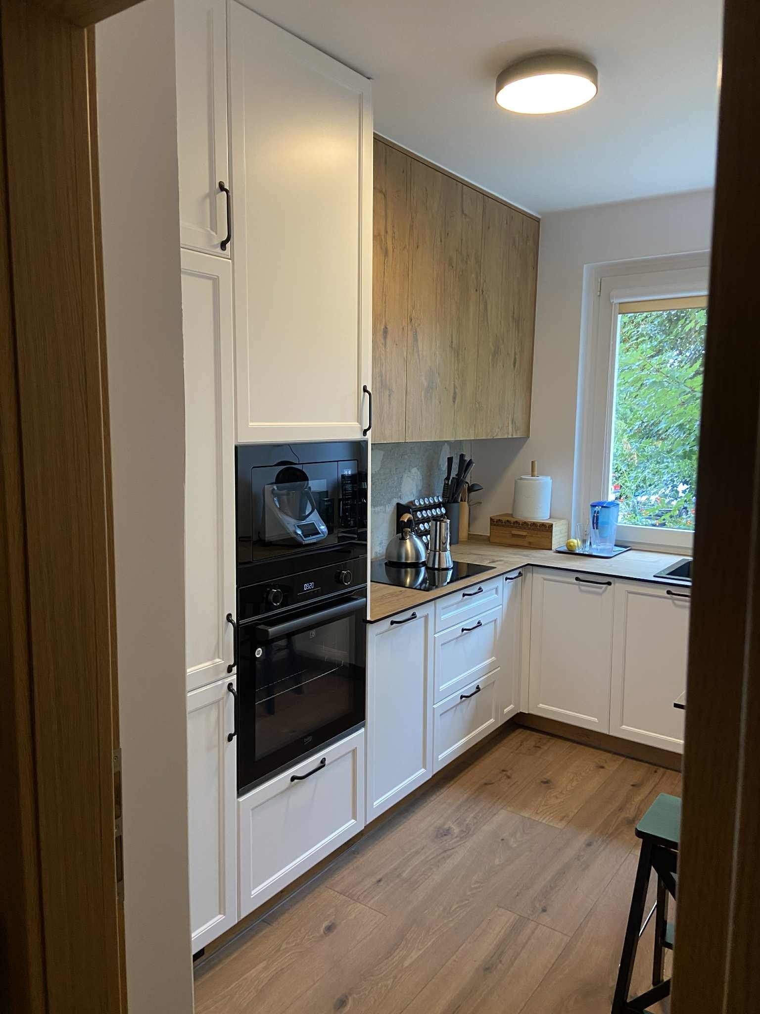 Nowoczesna kuchnia z białymi szafkami, czarnymi uchwytami i drewnianymi akcentami. Widoczny sprzęt AGD i drewniana podłoga. Ujęcie zza framugi drzwi.