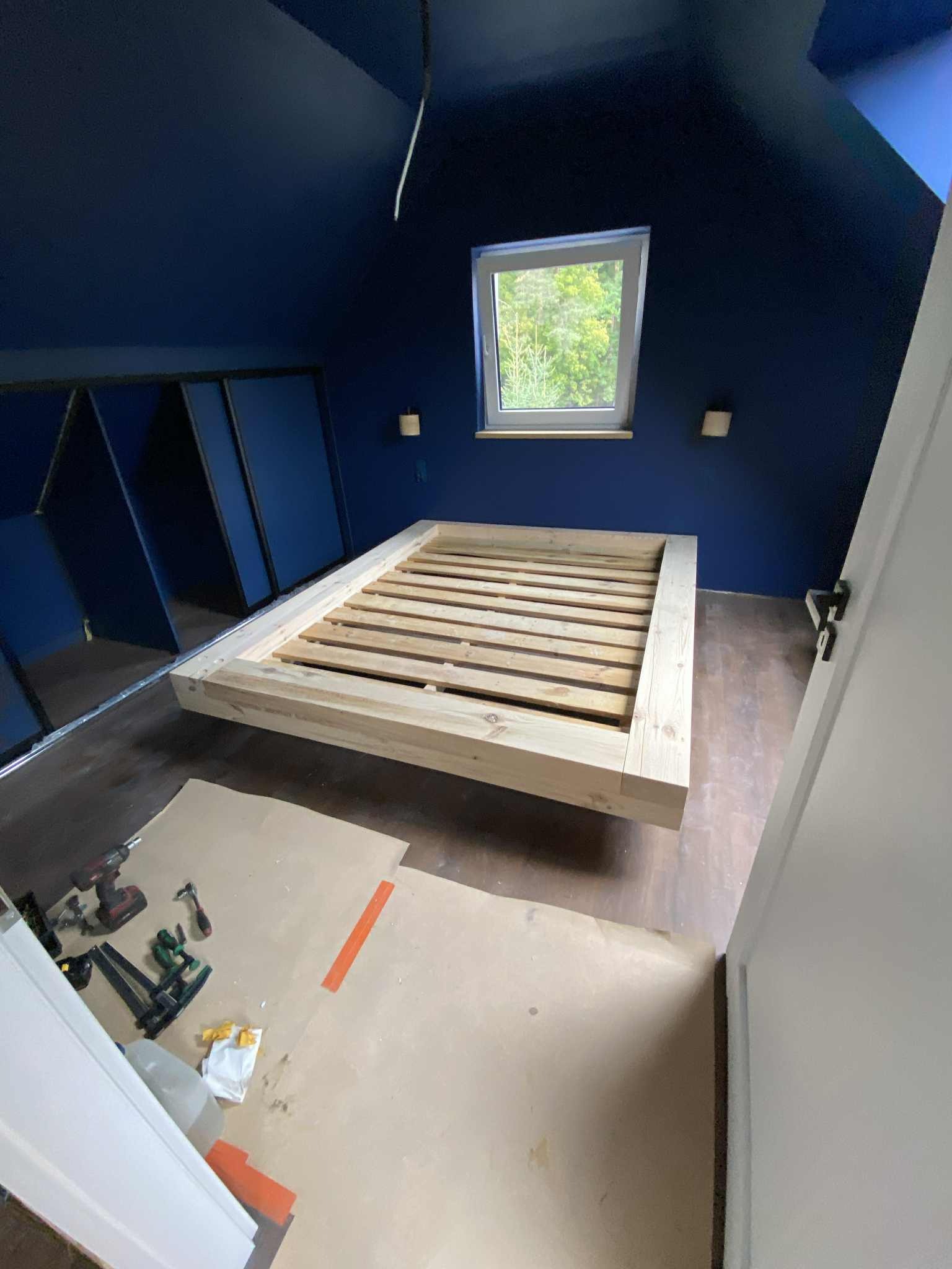 Surowa drewniana rama łóżka na poddaszu z niebieskimi ścianami, narzędzia stolarskie na podłodze. Widoczne okno i elementy konstrukcyjne szafy.