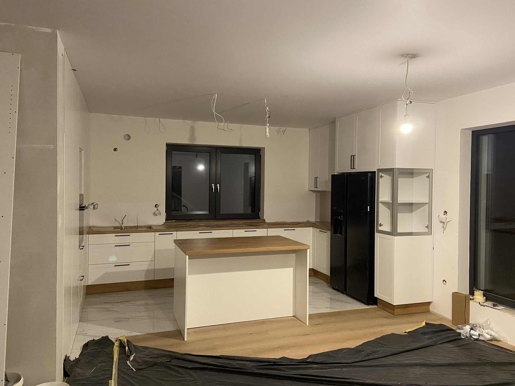 Wnętrze kuchni w trakcie montażu: białe szafki, drewniany blat, wyspa kuchenna. Na podłodze folia ochronna. Widoczne niedokończone instalacje elektryczne.