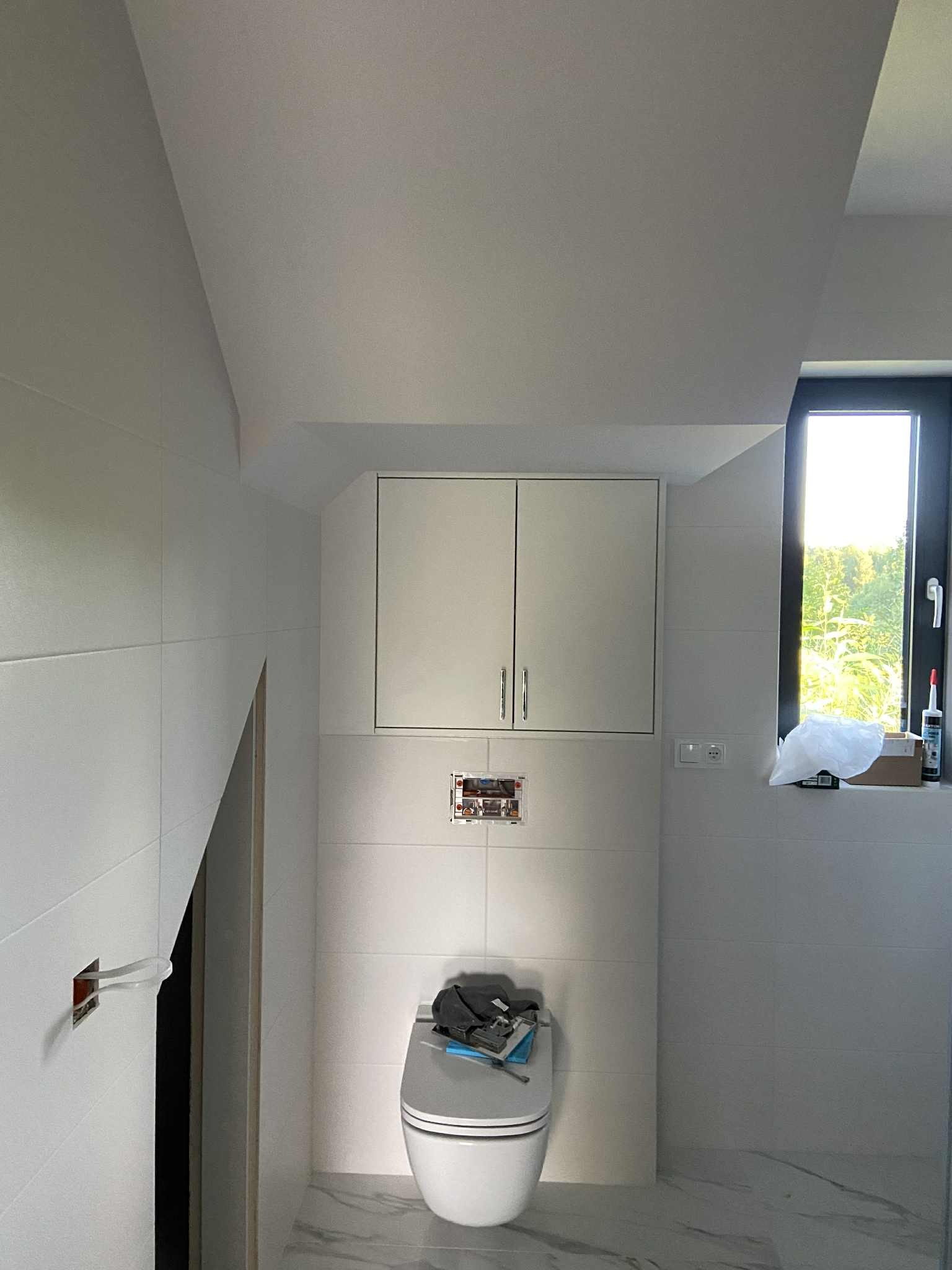 Biała łazienka w skosach z podwieszaną szafką nad toaletą. Widoczne płytki, okno i instalacja elektryczna w trakcie montażu. Podłoga imitująca marmur.