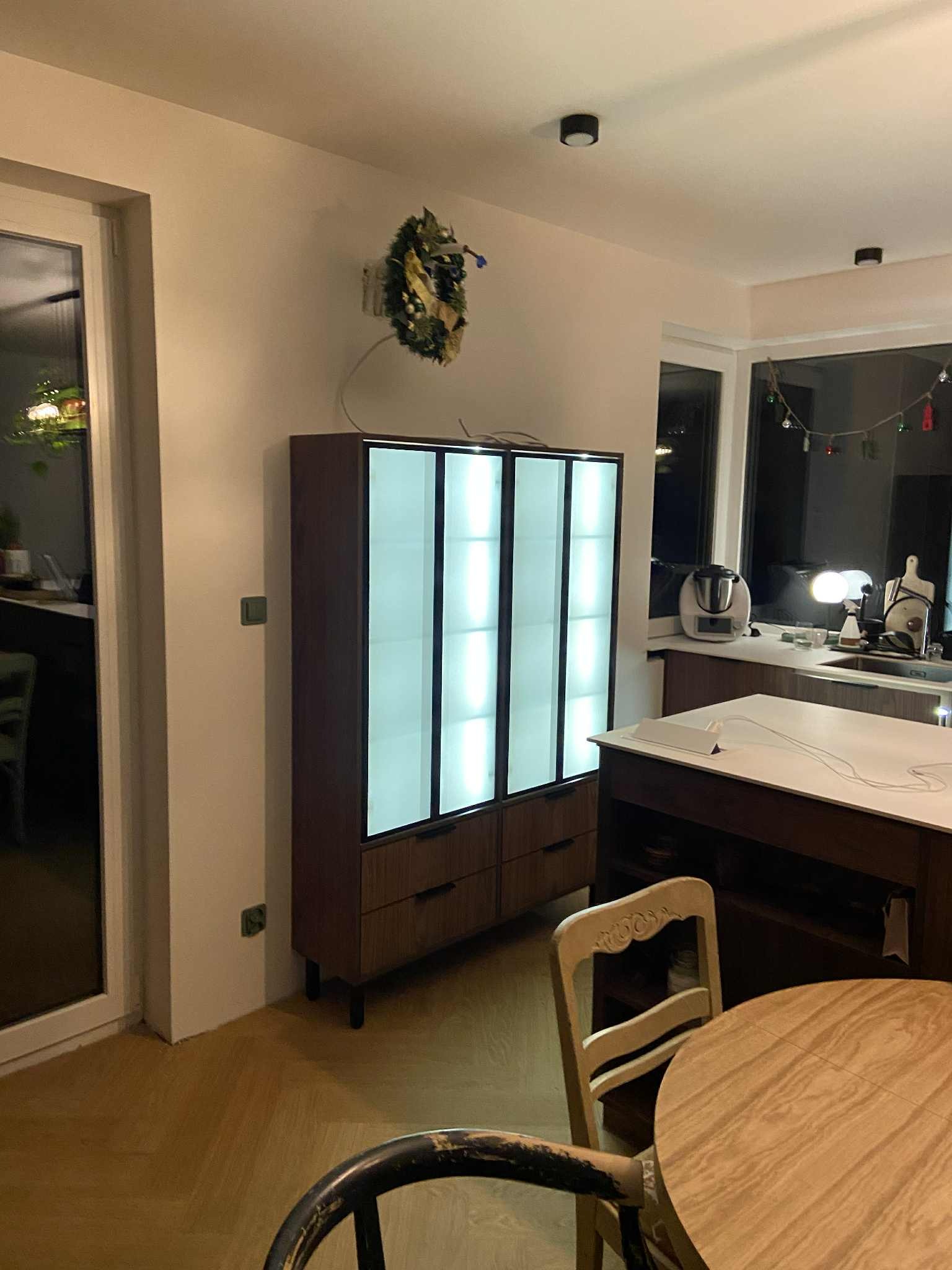 Nowoczesna szafa z podświetlanymi frontami w salonie, drewniane wykończenie, widoczny wieniec świąteczny, parkiet i fragment stołu z krzesłem.