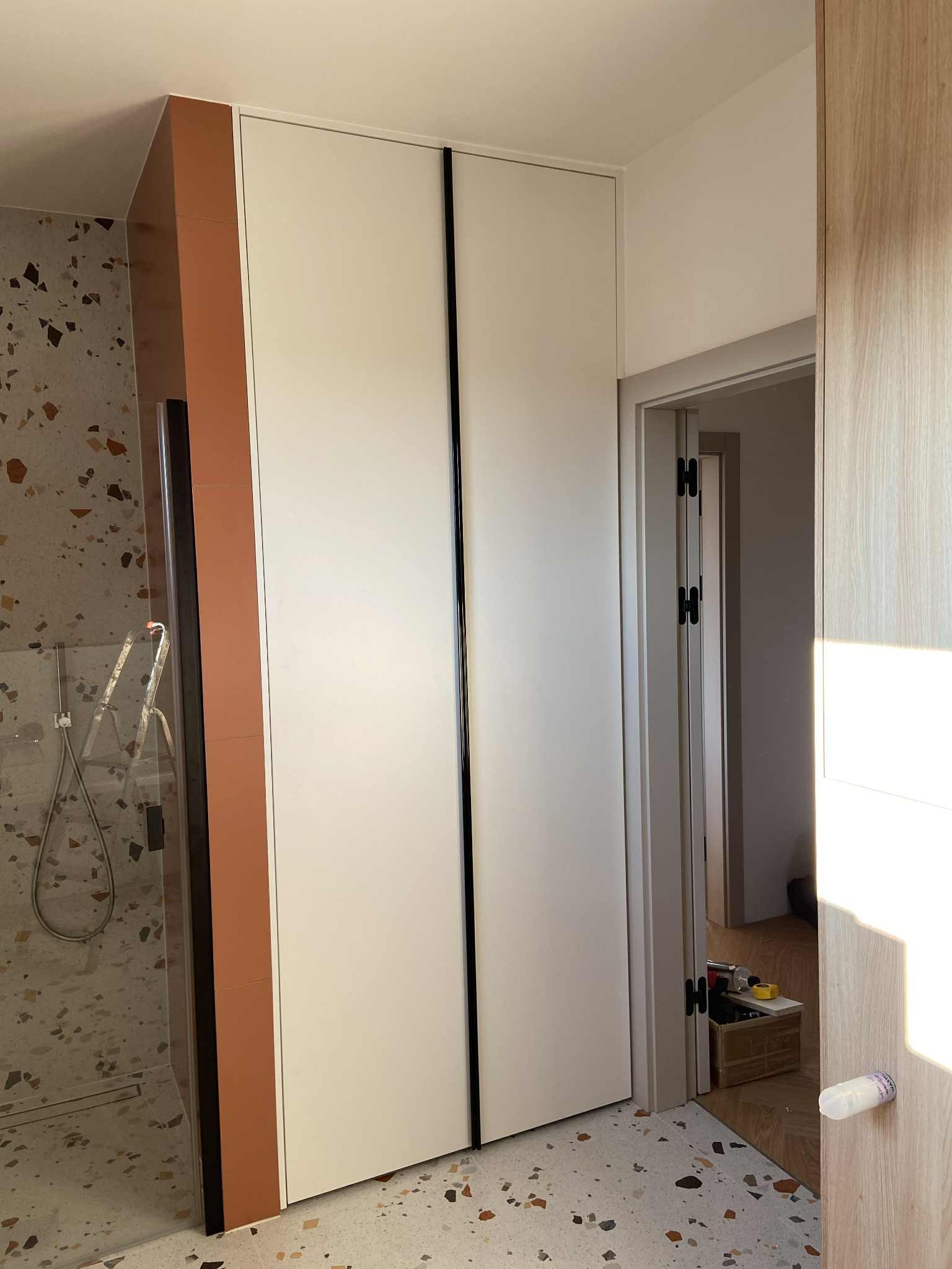 Biała szafa wnękowa z czarnymi uchwytami, w łazience z terazzo, widok na prysznic i otwarte drzwi do innego pomieszczenia. Szafa sięga od podłogi do sufitu.