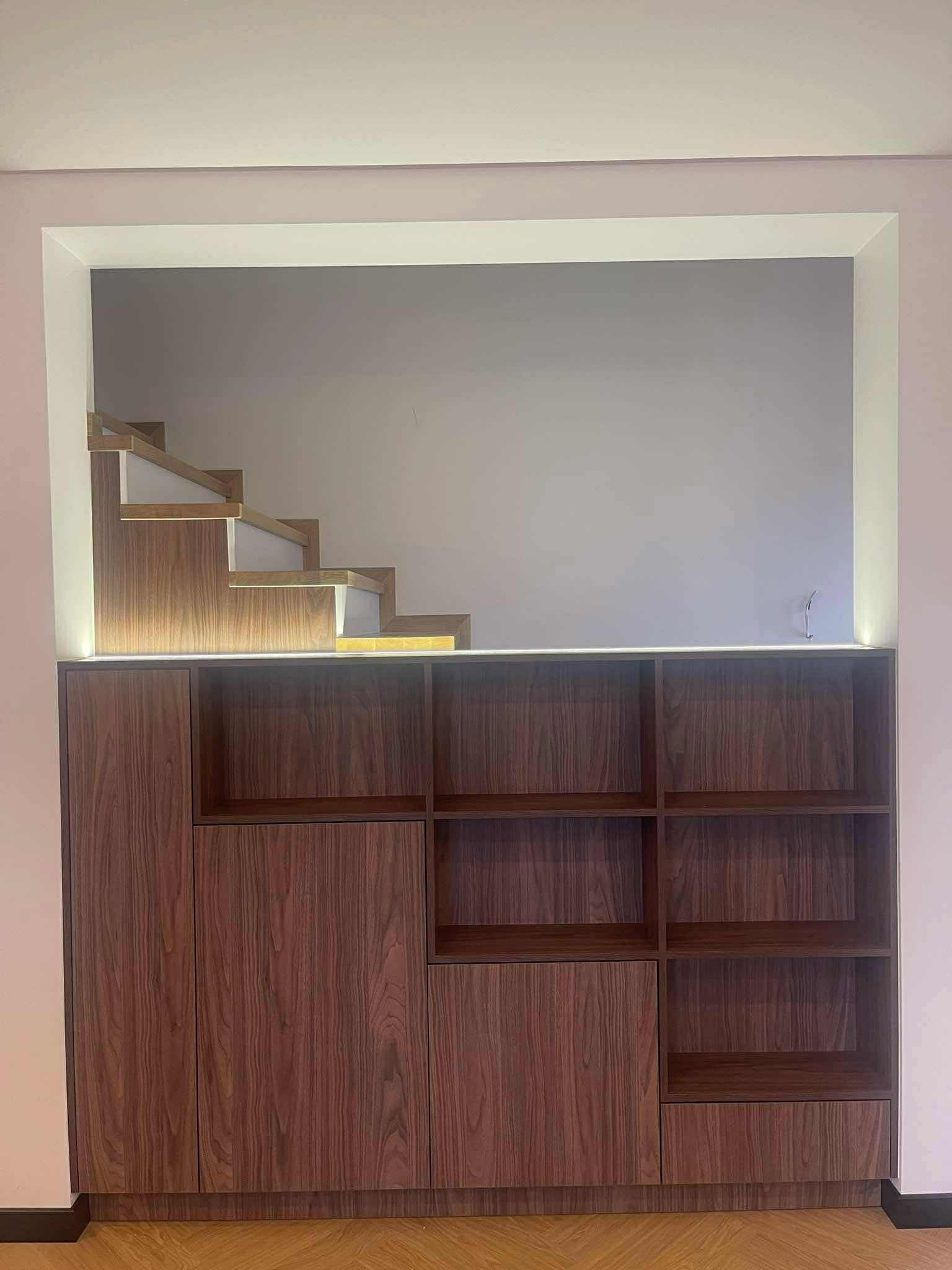 Nowoczesna szafa z drewna w kolorze orzecha, wbudowana we wnękę z podświetleniem LED, z widocznymi fragmentami schodów w górnej części. Minimalistyczny design.