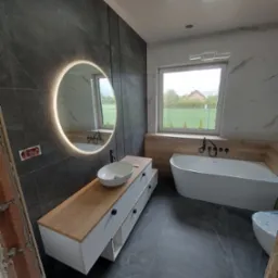 Nowoczesna łazienka z okrągłym lustrem z podświetleniem LED, białą wanną i umywalką nablatową. Ciemne płytki imitujące kamień na ścianach i podłodze.
