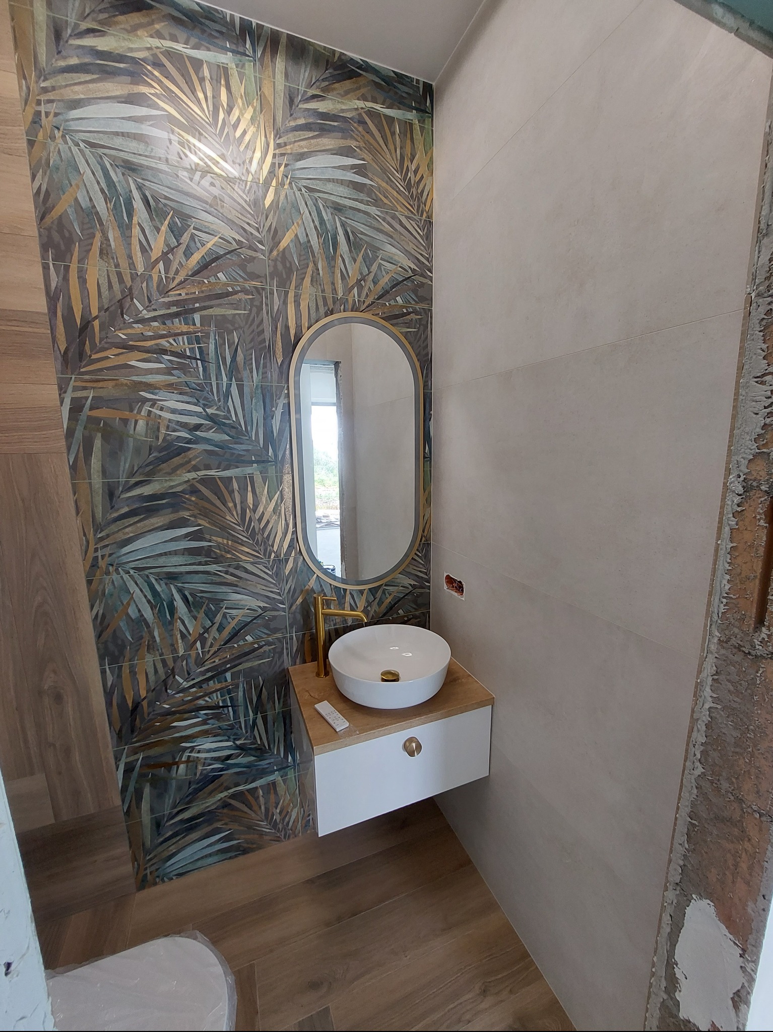 Nowoczesna łazienka po remoncie: umywalka nablatowa na szafce, lustro w złotej ramie i płytki z motywem liści palmowych na ścianie.