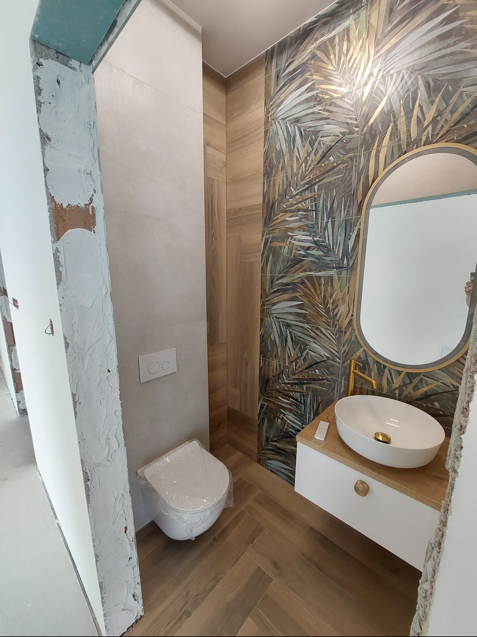 Nowoczesna łazienka w trakcie wykańczania: płytki z motywem liści, owalna umywalka na szafce z drewnianym blatem, toaleta w folii ochronnej, surowy fragment futryny.