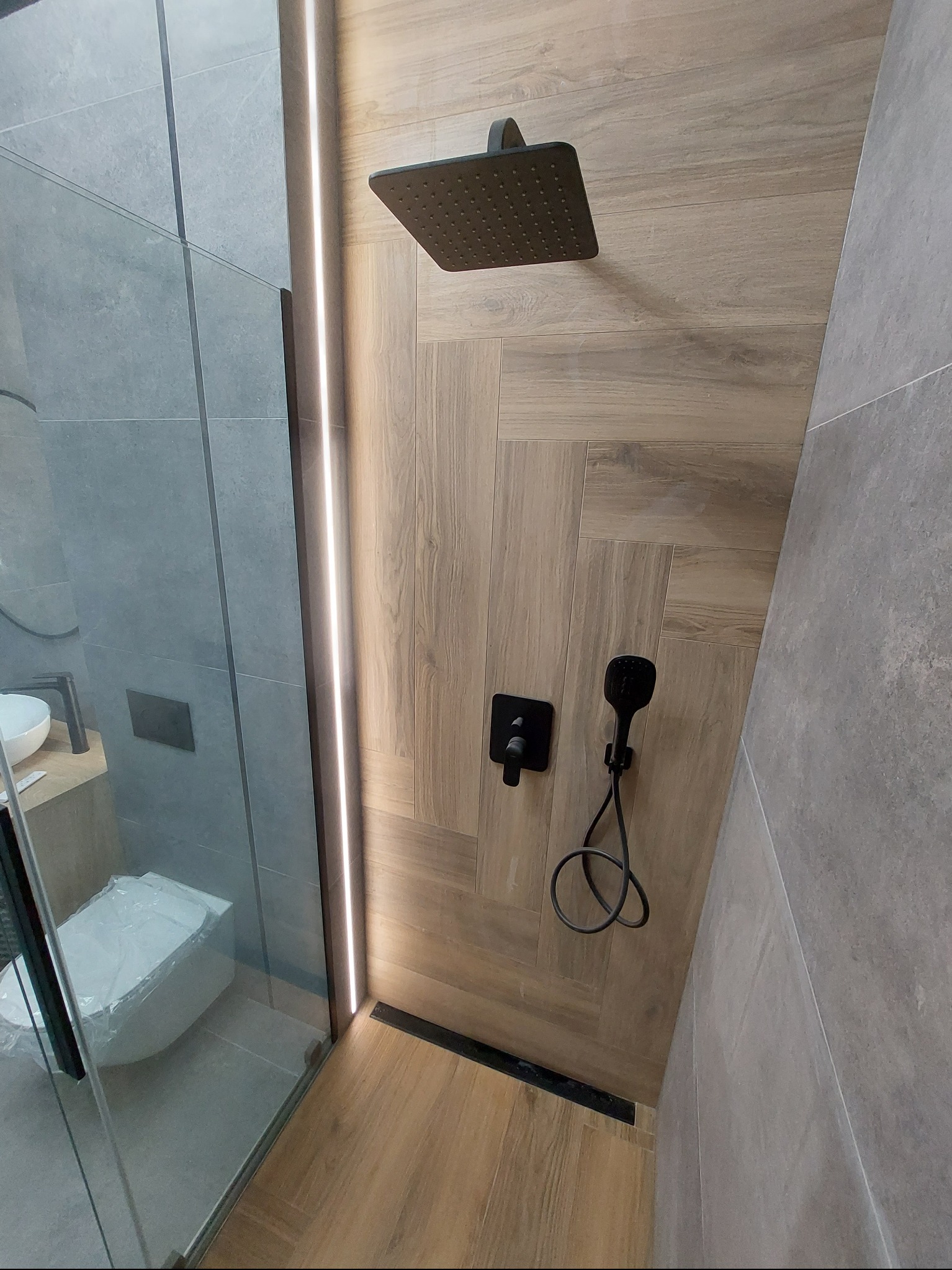 Nowoczesna łazienka z kabiną prysznicową typu walk-in, wykończona płytkami imitującymi drewno i szarymi płytami wielkoformatowymi. Czarna armatura i oświetlenie LED.