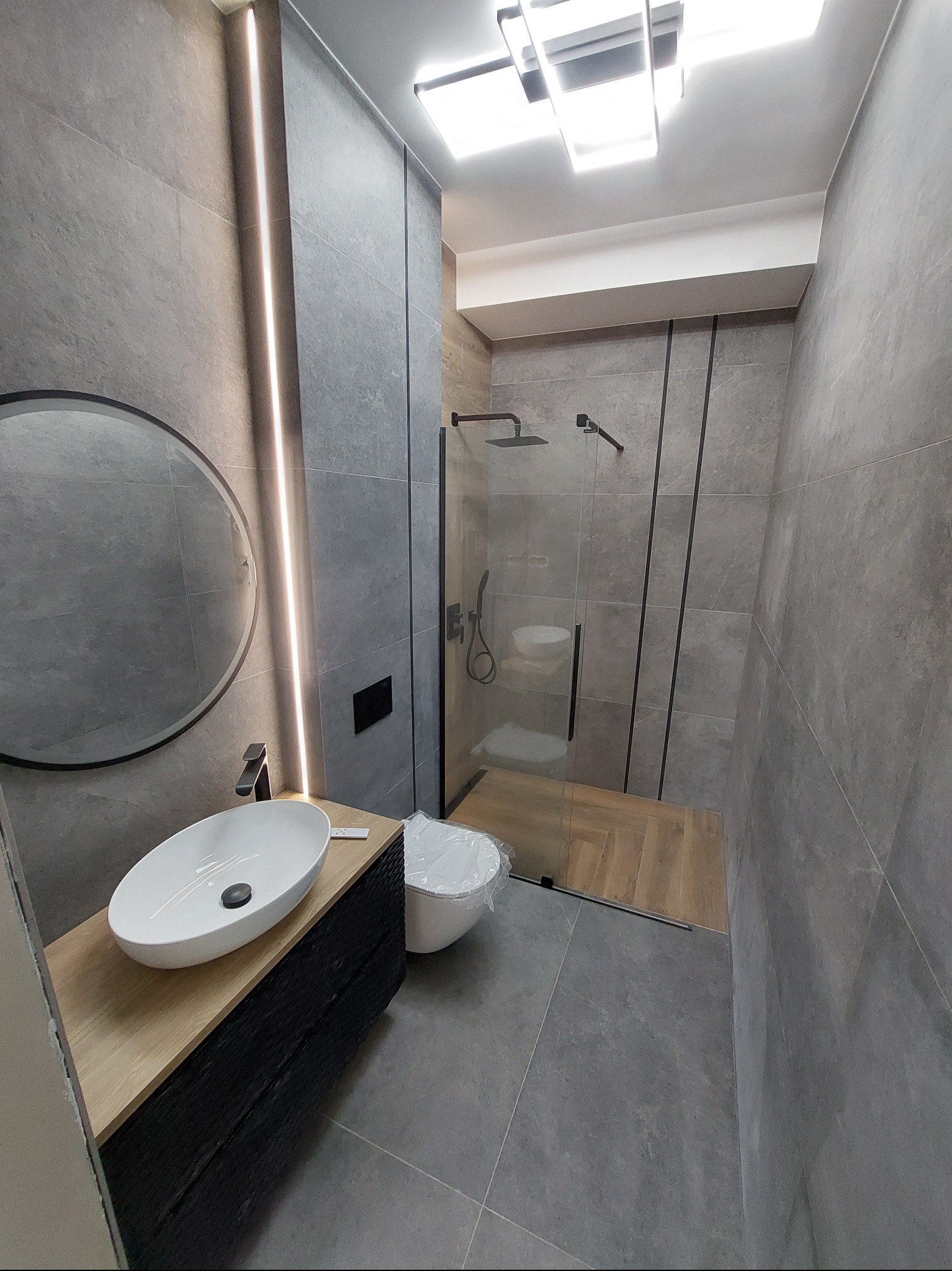 Stylowa łazienka w szarościach z okrągłym lustrem, podświetleniem LED i kabiną prysznicową walk-in. Umywalka nablatowa na drewnianej szafce. Minimalistyczny design.