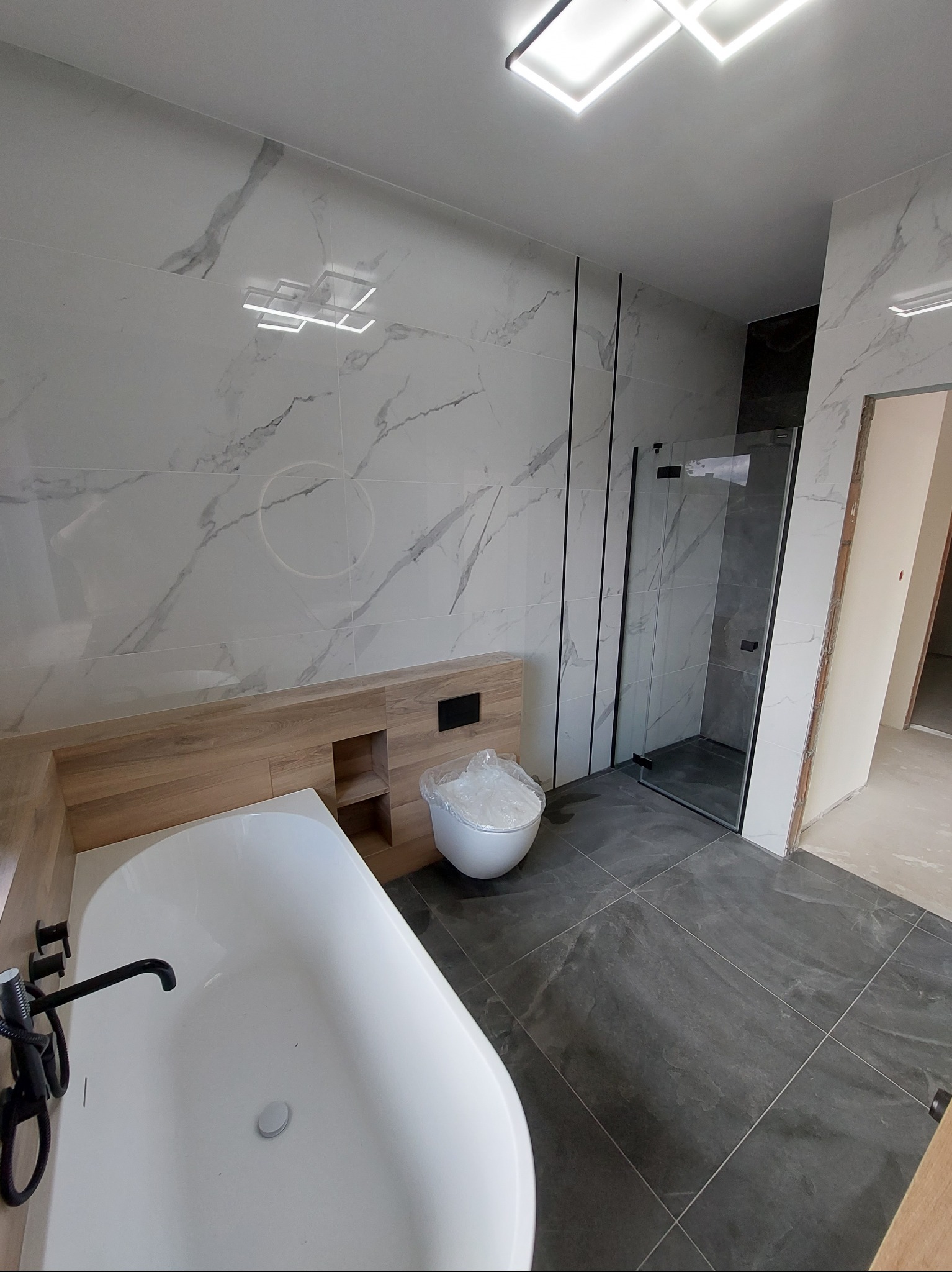 Nowoczesna łazienka po remoncie: biała wanna z czarną armaturą, szare płytki podłogowe, białe marmurowe płytki ścienne i kabina prysznicowa ze szklanymi drzwiami.