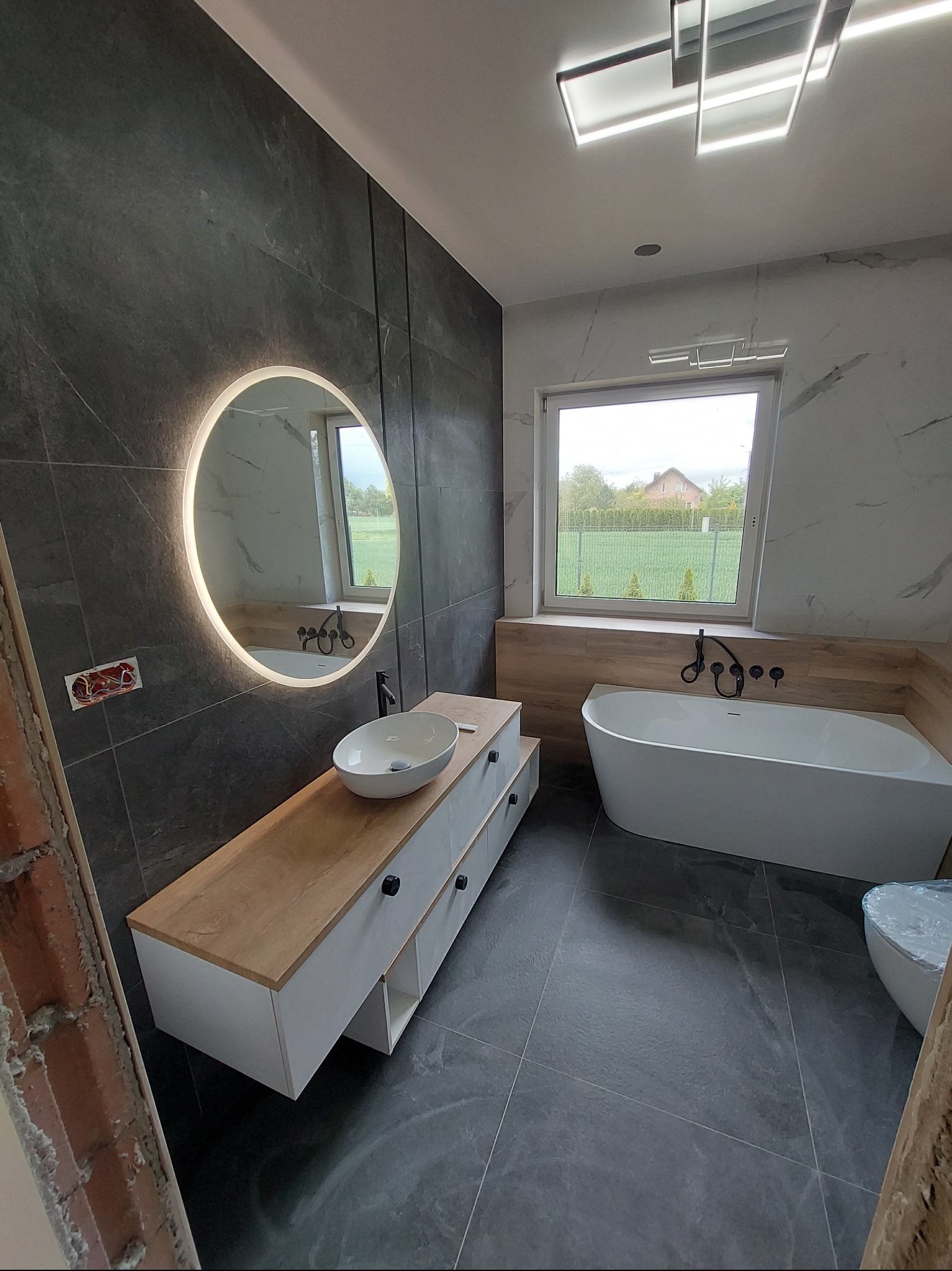 Nowoczesna łazienka z okrągłym lustrem z podświetleniem LED, białą wanną i umywalką nablatową. Ciemne płytki imitujące kamień na ścianach i podłodze.