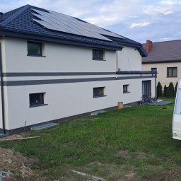 OGGITECH SPÓŁKA Z OGRANICZONĄ ODPOWIEDZIALNOŚCIĄ - Dwukondygnacyjny dom z panelami słonecznymi na dachu, świeżo pomalowaną fasadą w odcieniach bieli i szarości, otoczony trawnikiem i częściowo ułożoną kostką.