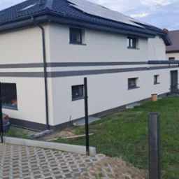 Dwukondygnacyjny dom z panelami słonecznymi na dachu, świeżo pomalowany na biało z szarymi akcentami, widoczny z perspektywy trawnika i częściowo wyłożonego kostką podjazdu.