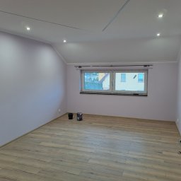 OGGITECH SPÓŁKA Z OGRANICZONĄ ODPOWIEDZIALNOŚCIĄ - Pomalowane na liliowo poddasze z drewnopodobną podłogą i oknem. Widoczne puszki z farbą. Oświetlenie sufitowe punktowe. Częściowo widoczne drewniane drzwi.