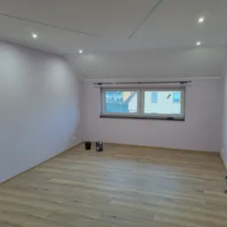 Pomalowane na liliowo poddasze z drewnopodobną podłogą i oknem. Widoczne puszki z farbą. Oświetlenie sufitowe punktowe. Częściowo widoczne drewniane drzwi.