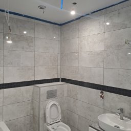 OGGITECH SPÓŁKA Z OGRANICZONĄ ODPOWIEDZIALNOŚCIĄ - Wykończona łazienka z beżowymi płytkami, czarnym pasem dekoracyjnym, białą umywalką i toaletą. Widoczne niedokończone elementy instalacji elektrycznej.