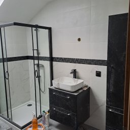 OGGITECH SPÓŁKA Z OGRANICZONĄ ODPOWIEDZIALNOŚCIĄ - Nowoczesna łazienka w trakcie wykończenia: kabina prysznicowa z czarną ramą, umywalka nablatowa, biało-czarne płytki, widoczne narzędzia i materiały budowlane.