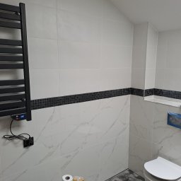 OGGITECH SPÓŁKA Z OGRANICZONĄ ODPOWIEDZIALNOŚCIĄ - Wykończona łazienka z białymi płytkami imitującymi marmur, czarnym pasem mozaiki i grzejnikiem drabinkowym. Widoczna toaleta i akcesoria łazienkowe.