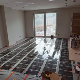 OGGITECH SPÓŁKA Z OGRANICZONĄ ODPOWIEDZIALNOŚCIĄ - Przygotowanie do instalacji ogrzewania podłogowego: rozłożone maty grzewcze na podłożu w jasnym pokoju z dużym oknem balkonowym, widoczne narzędzia i materiały.