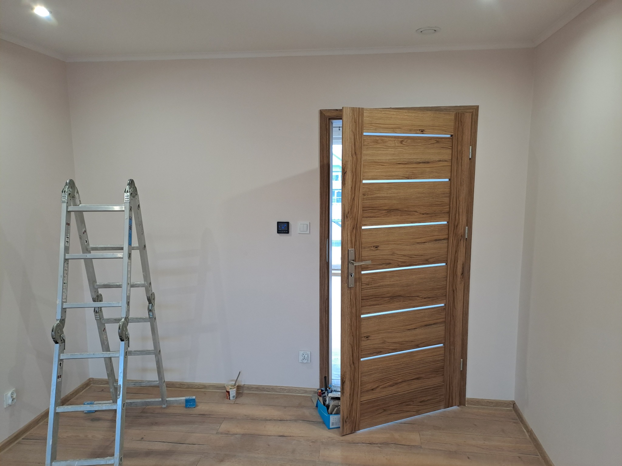 Otwarta, drewniana, nowoczesna drzwi wewnętrzne z poziomymi, białymi paskami. Ściana świeżo pomalowana na jasny kolor. Aluminiowa drabina obok. Podłoga drewnopodobna.
