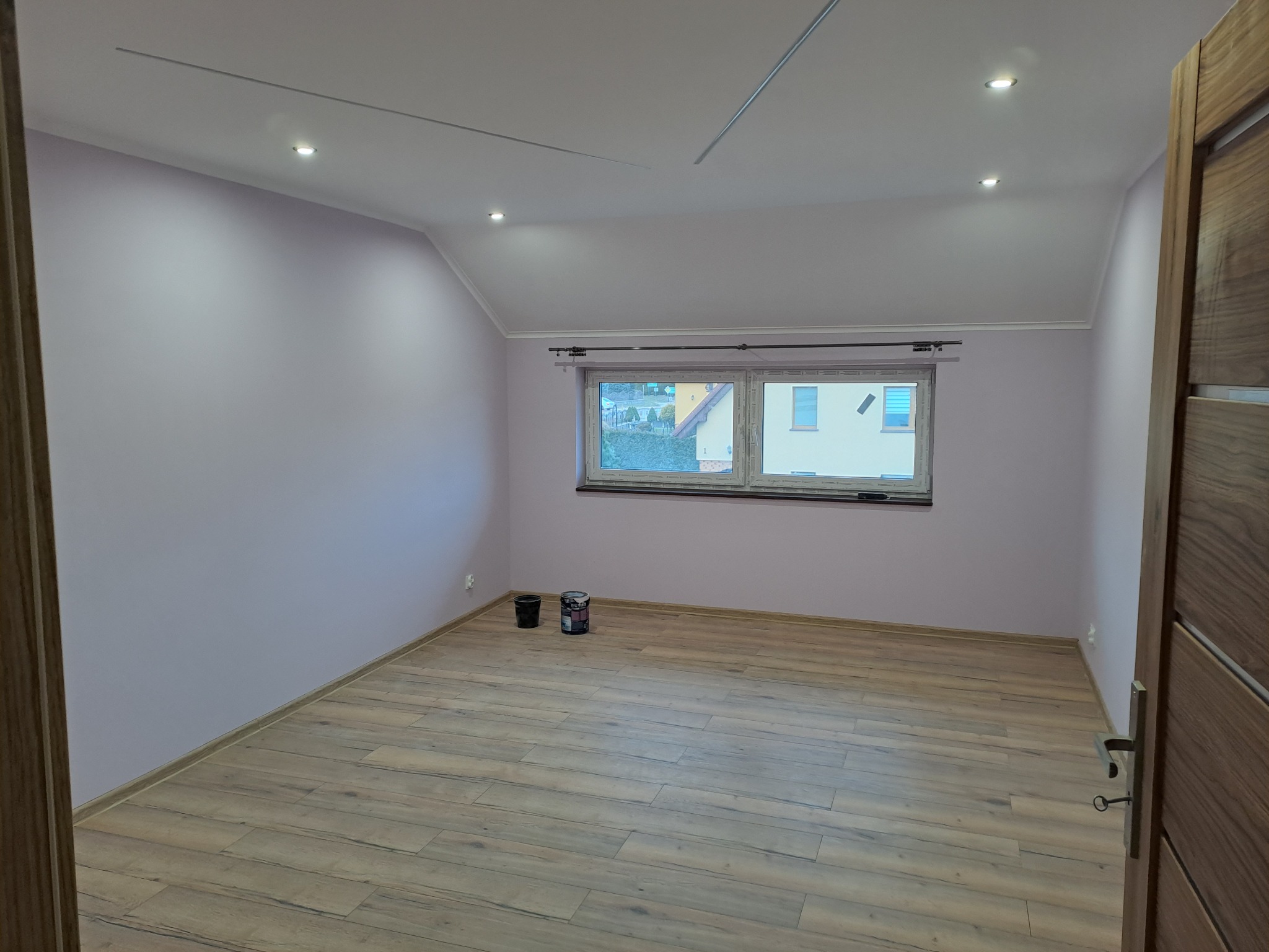 Pomalowane na liliowo poddasze z drewnopodobną podłogą i oknem. Widoczne puszki z farbą. Oświetlenie sufitowe punktowe. Częściowo widoczne drewniane drzwi.