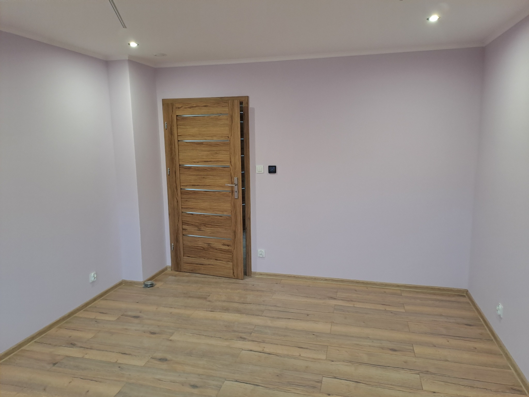 Jasne wnętrze po remoncie: drewniana podłoga, drzwi z poziomymi panelami, świeżo pomalowane ściany w odcieniach fioletu, widoczne gniazdka i oświetlenie punktowe.