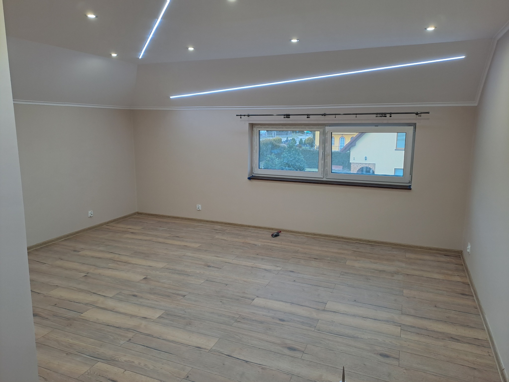 Jasne panele podłogowe ułożone w pustym pokoju z oknem i nowoczesnym oświetleniem LED w Gościcinie. Kremowe ściany, czyste wykończenie.