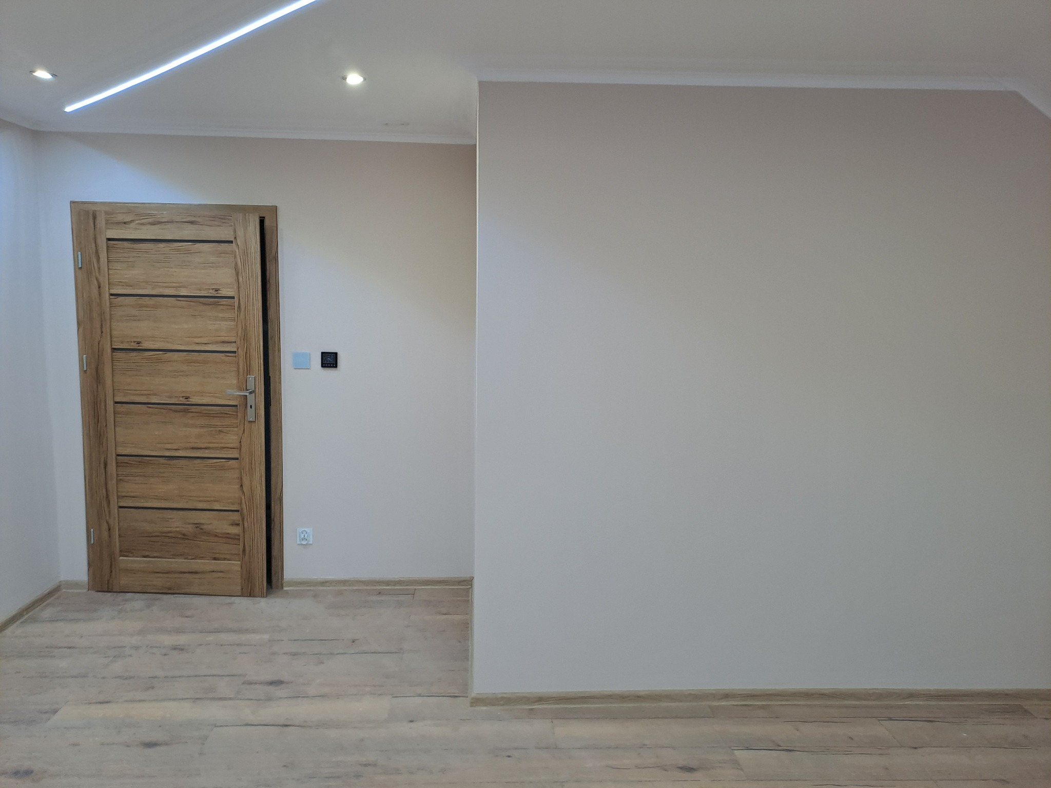 Jasne wnętrze po remoncie: drewniane drzwi z poziomymi panelami, parkiet w odcieniach beżu i szarości, świeżo pomalowane ściany w neutralnym odcieniu, nowoczesne oświetlenie.