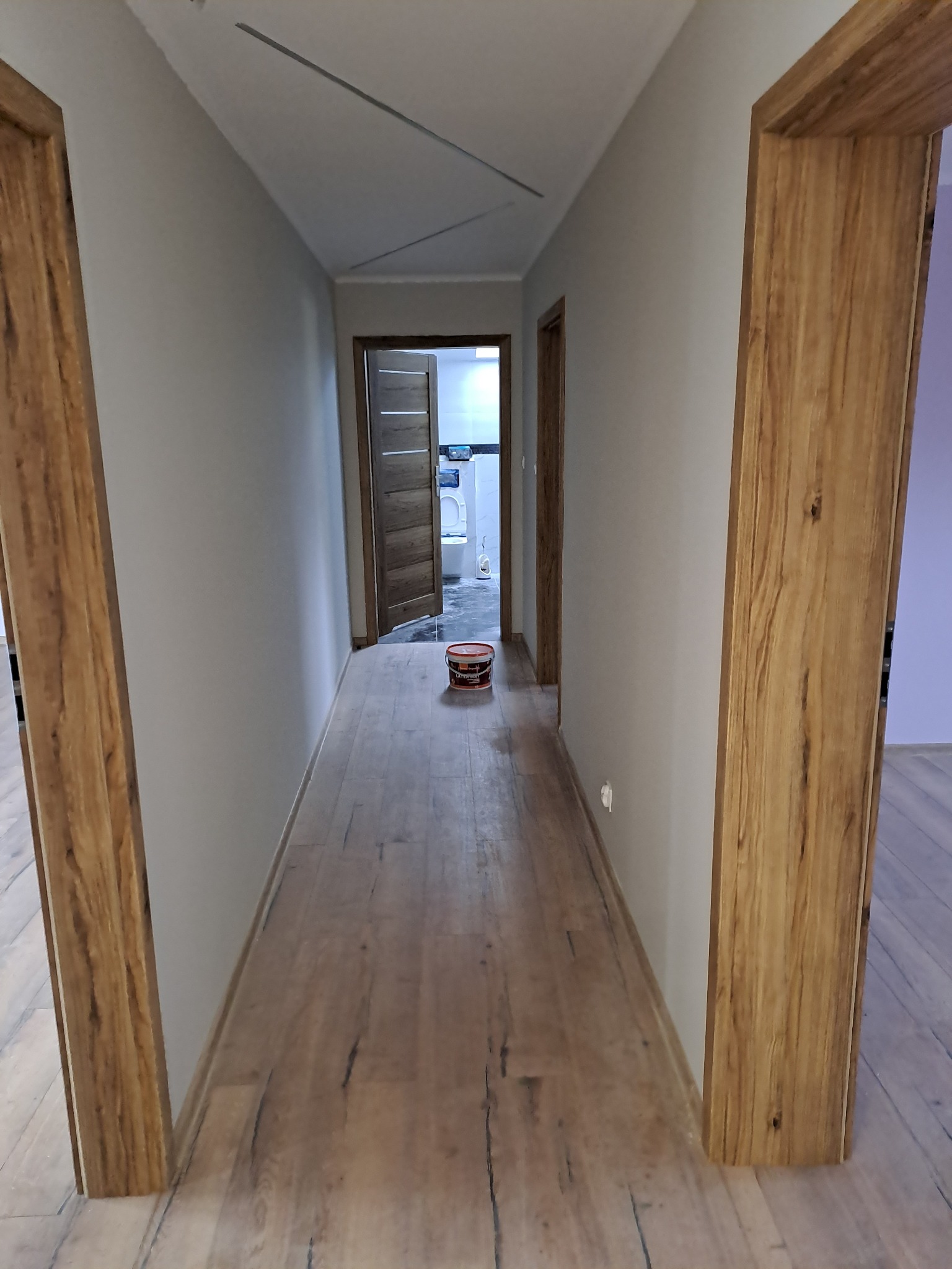 Korytarz po remoncie: świeżo pomalowane ściany w odcieniach szarości, drewnopodobna podłoga i drzwi z ościeżnicami o fakturze drewna. Widoczna puszka z farbą.