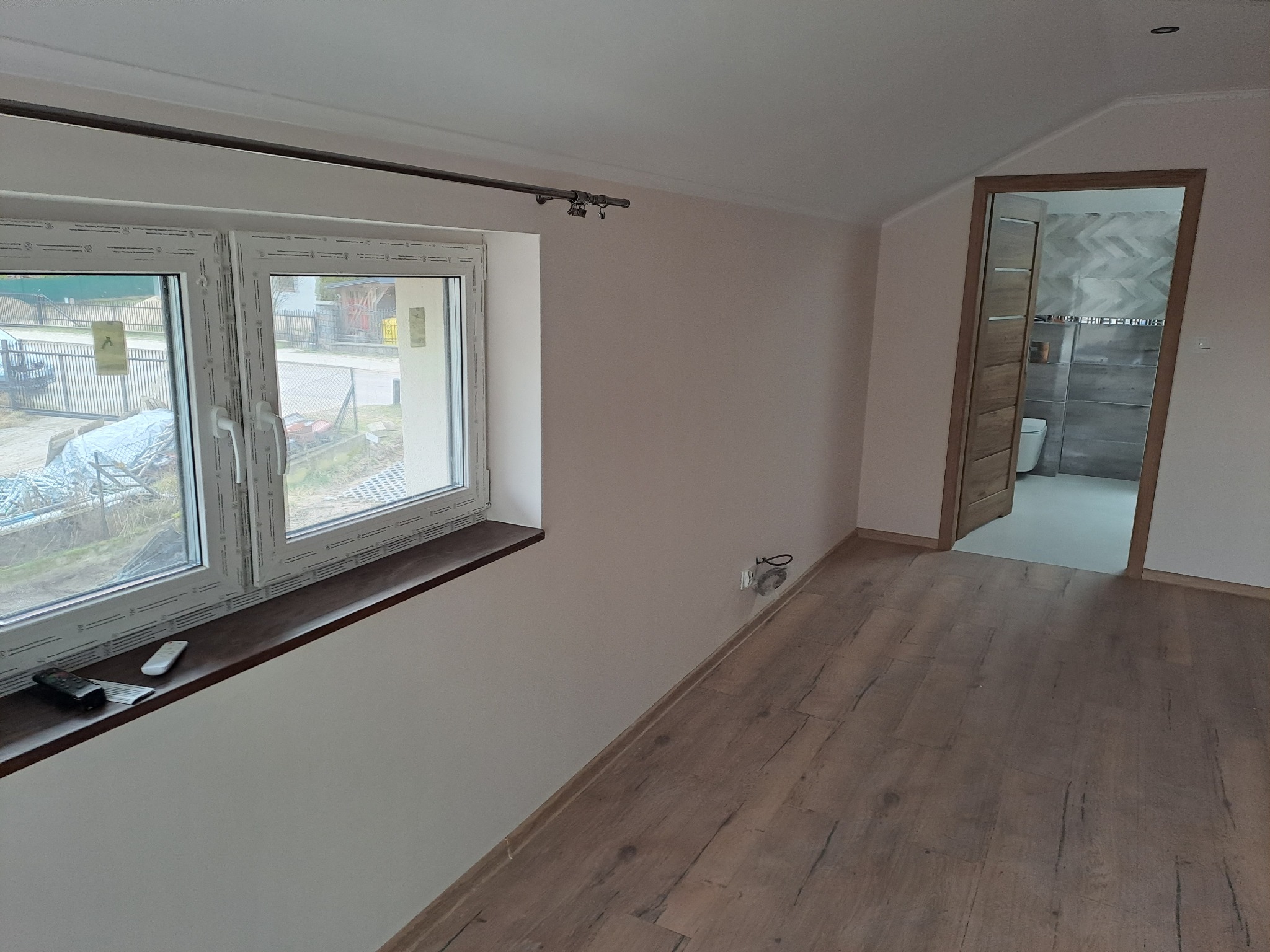 Jasne wnętrze z oknem i drewnopodobną podłogą. W tle widoczna łazienka z szarą, wzorzystą glazurą. Na parapecie leżą piloty. Świeżo pomalowane ściany.