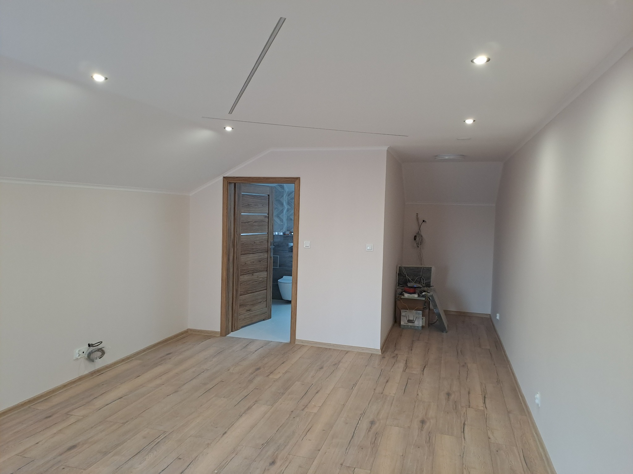 Jasne, przestronne wnętrze po remoncie: drewnopodobna podłoga, świeżo pomalowane ściany w odcieniach bieli, otwarte drzwi do łazienki z widoczną armaturą.