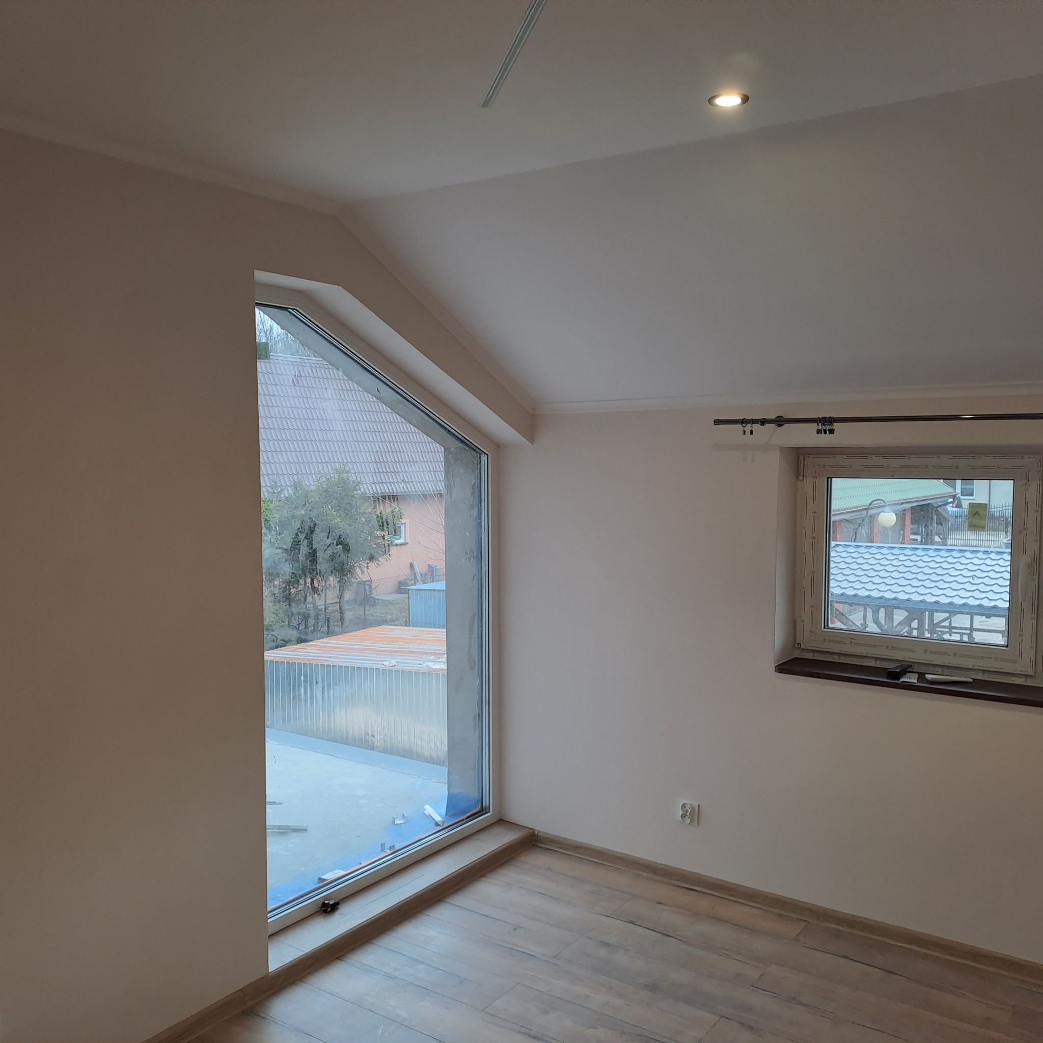 Jasne wnętrze poddasza po remoncie: ściany pomalowane na beżowo, parkiet, okno dachowe z widokiem na zewnątrz, okno standardowe z roletą. Gniazdko elektryczne widoczne na ścianie.