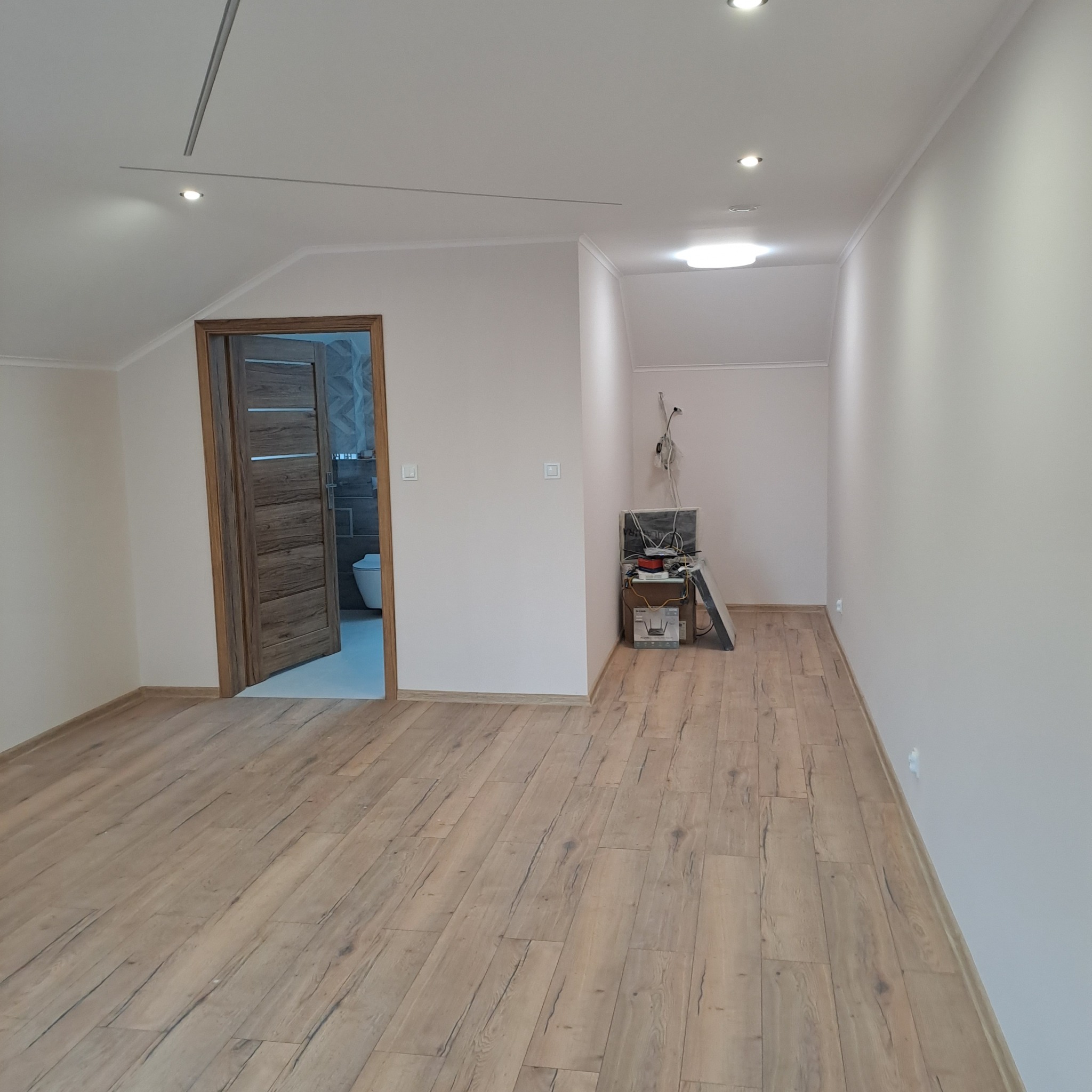 Jasne drewniane panele i świeżo pomalowane ściany w pokoju na poddaszu. W tle otwarte drzwi do łazienki z widoczną toaletą i szarą glazurą.