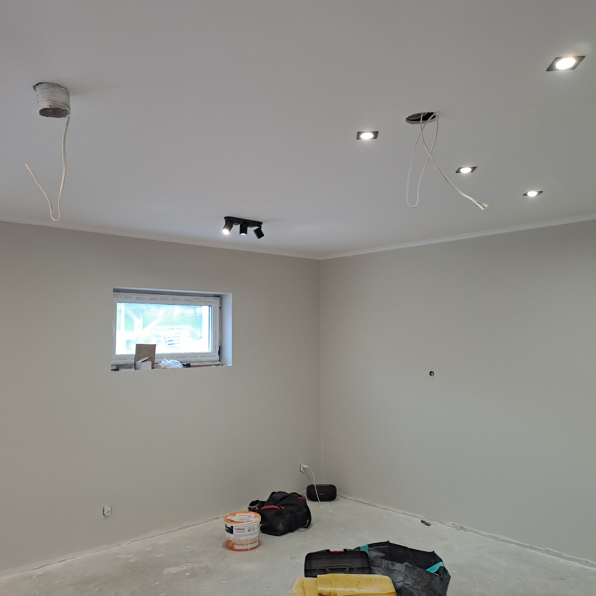 Szpachlowana ściana w jasnym odcieniu szarości z oknem i zamontowanym oświetleniem sufitowym. Widoczne narzędzia malarskie i puszka farby.