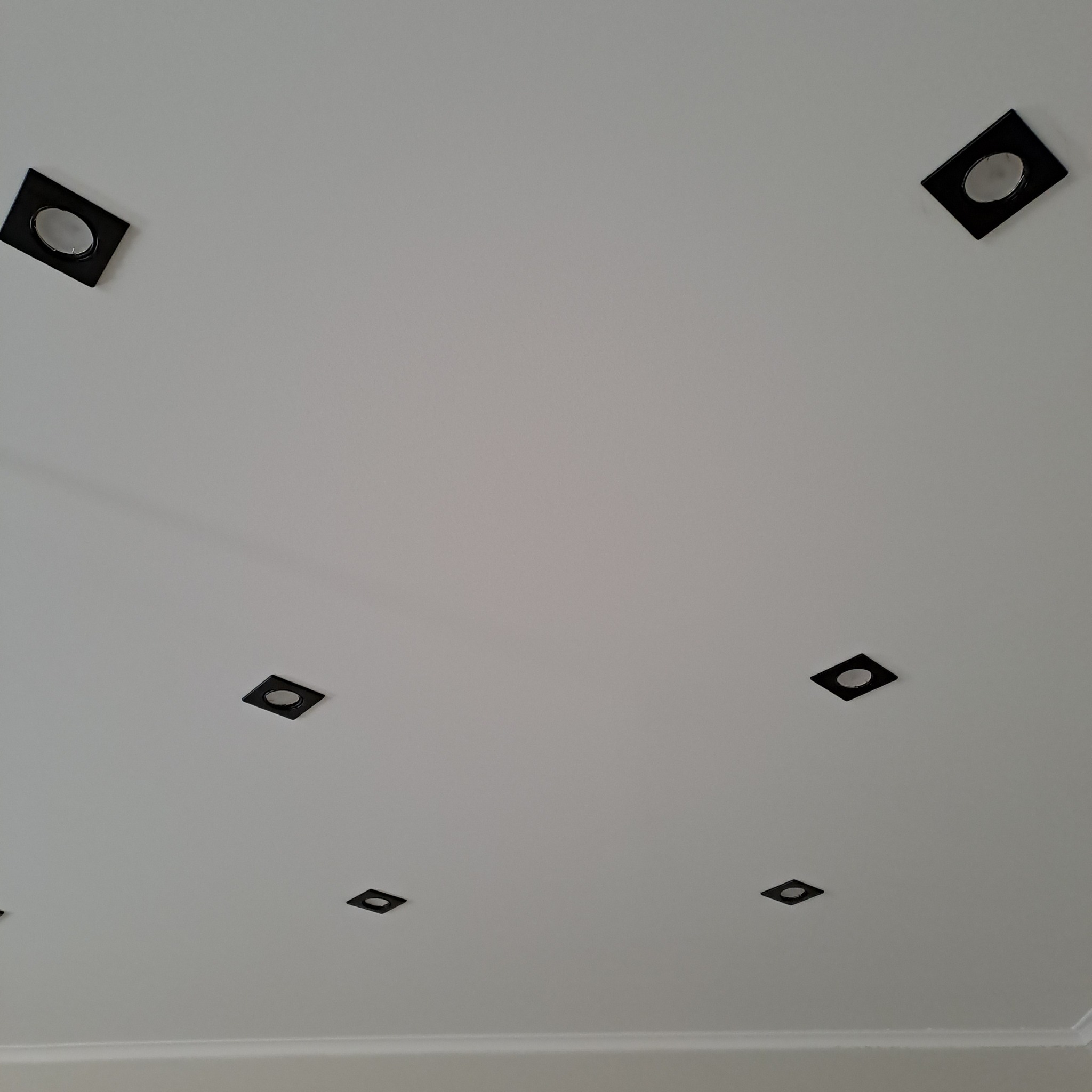 Biały sufit z równomiernie rozmieszczonymi, kwadratowymi, czarnymi oprawami oświetleniowymi typu downlight, tworzącymi nowoczesny wzór.