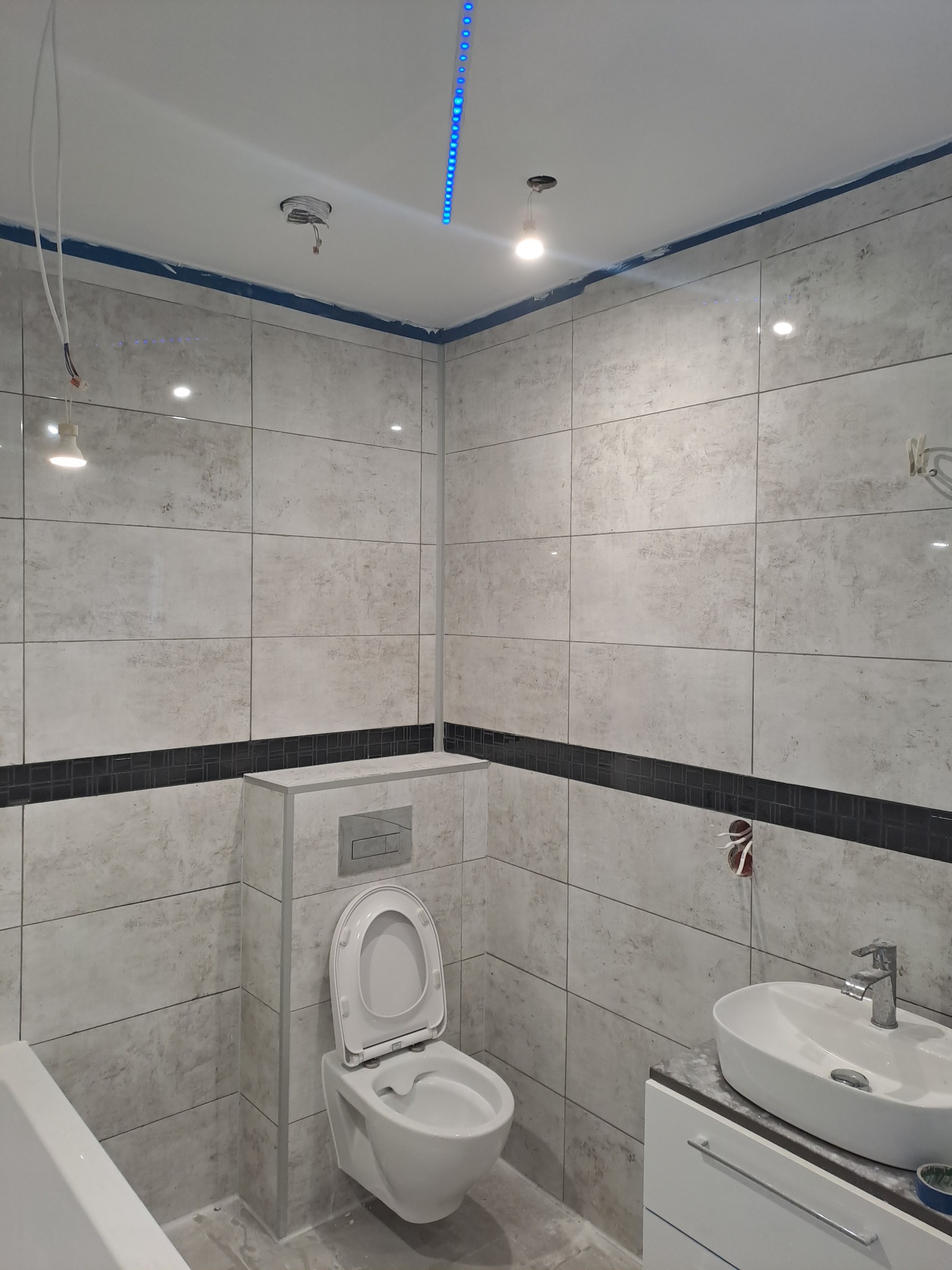 Wykończona łazienka z beżowymi płytkami, czarnym pasem dekoracyjnym, białą umywalką i toaletą. Widoczne niedokończone elementy instalacji elektrycznej.