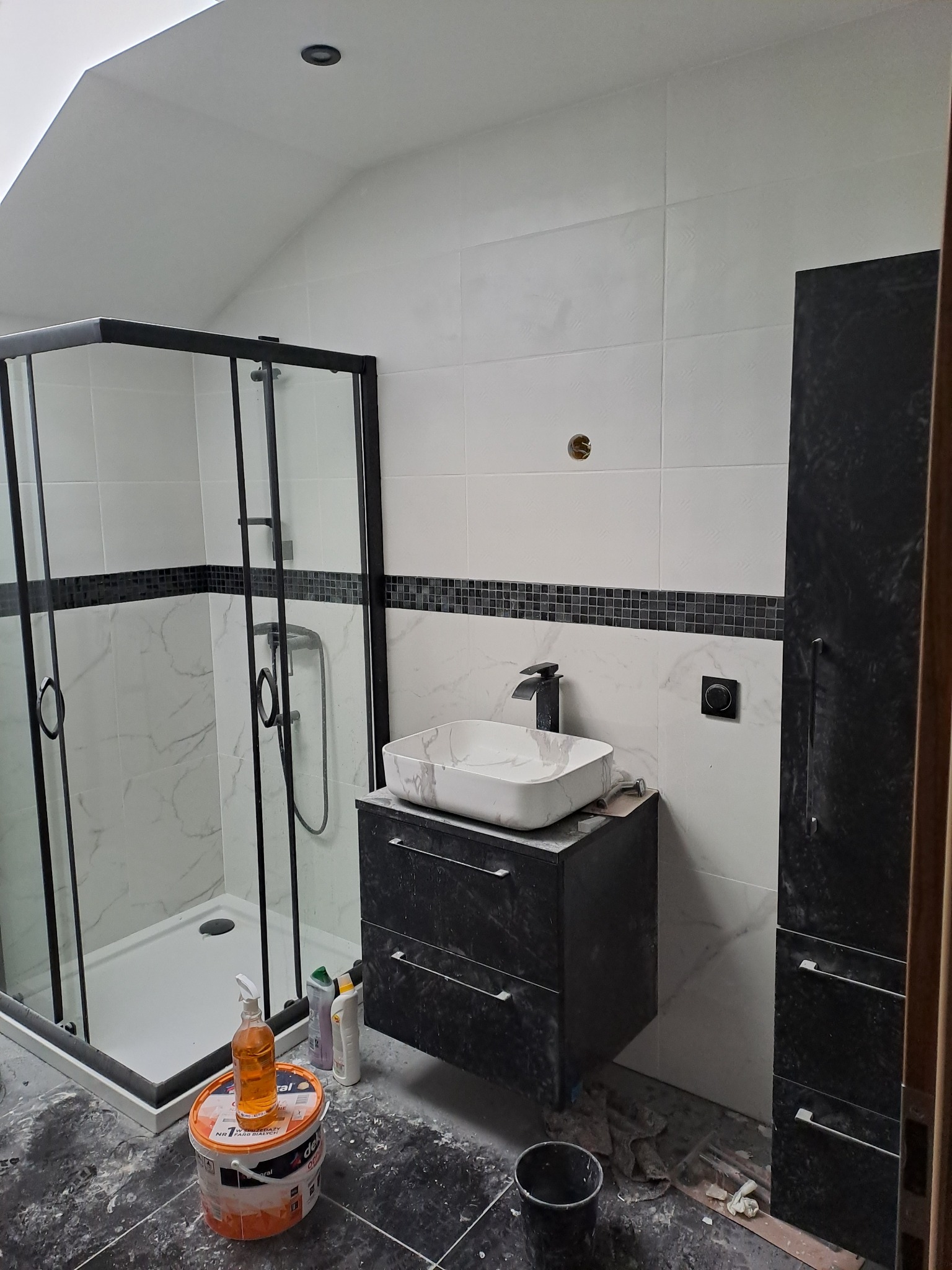 Nowoczesna łazienka w trakcie wykończenia: kabina prysznicowa z czarną ramą, umywalka nablatowa, biało-czarne płytki, widoczne narzędzia i materiały budowlane.