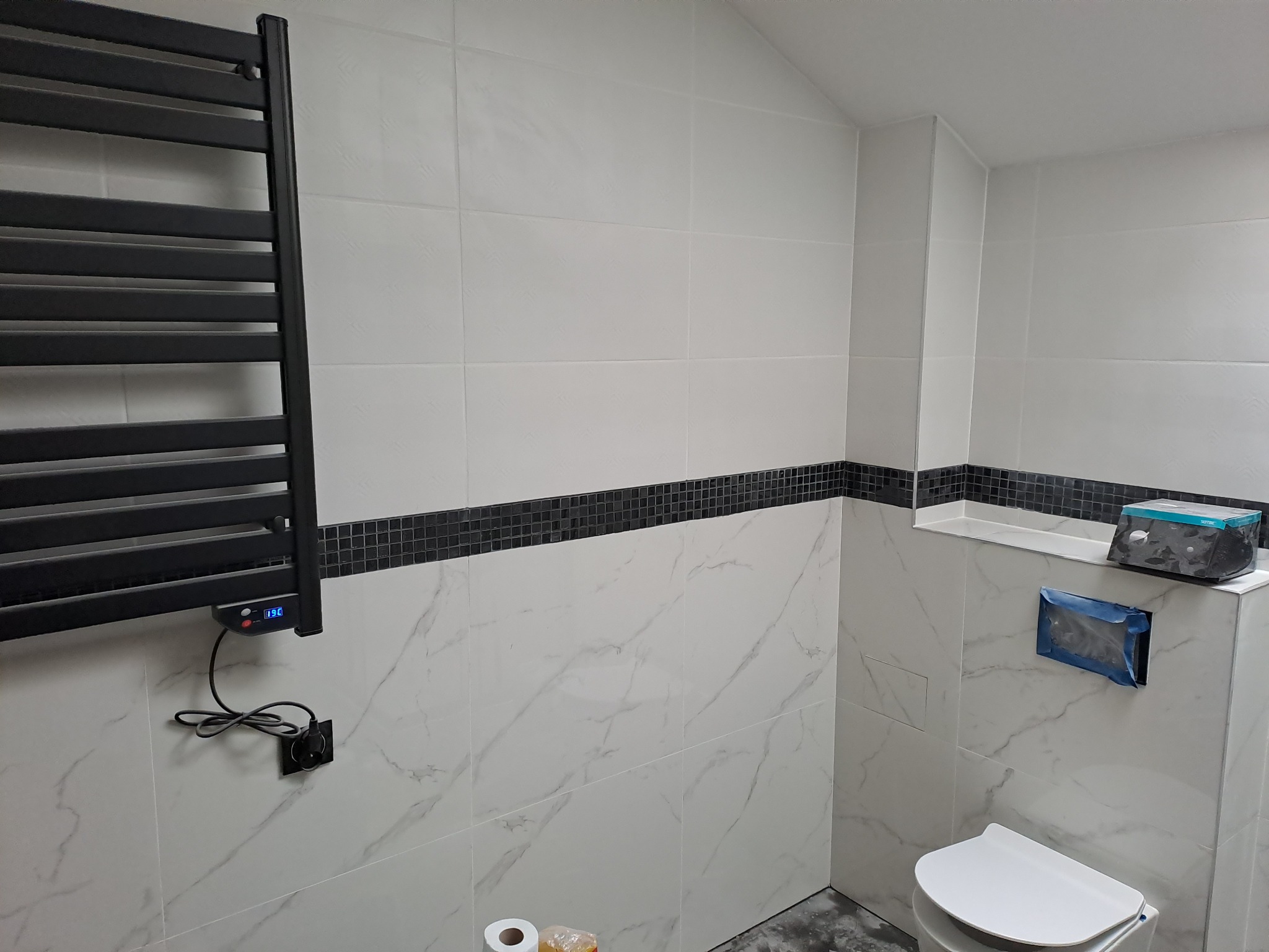 Wykończona łazienka z białymi płytkami imitującymi marmur, czarnym pasem mozaiki i grzejnikiem drabinkowym. Widoczna toaleta i akcesoria łazienkowe.