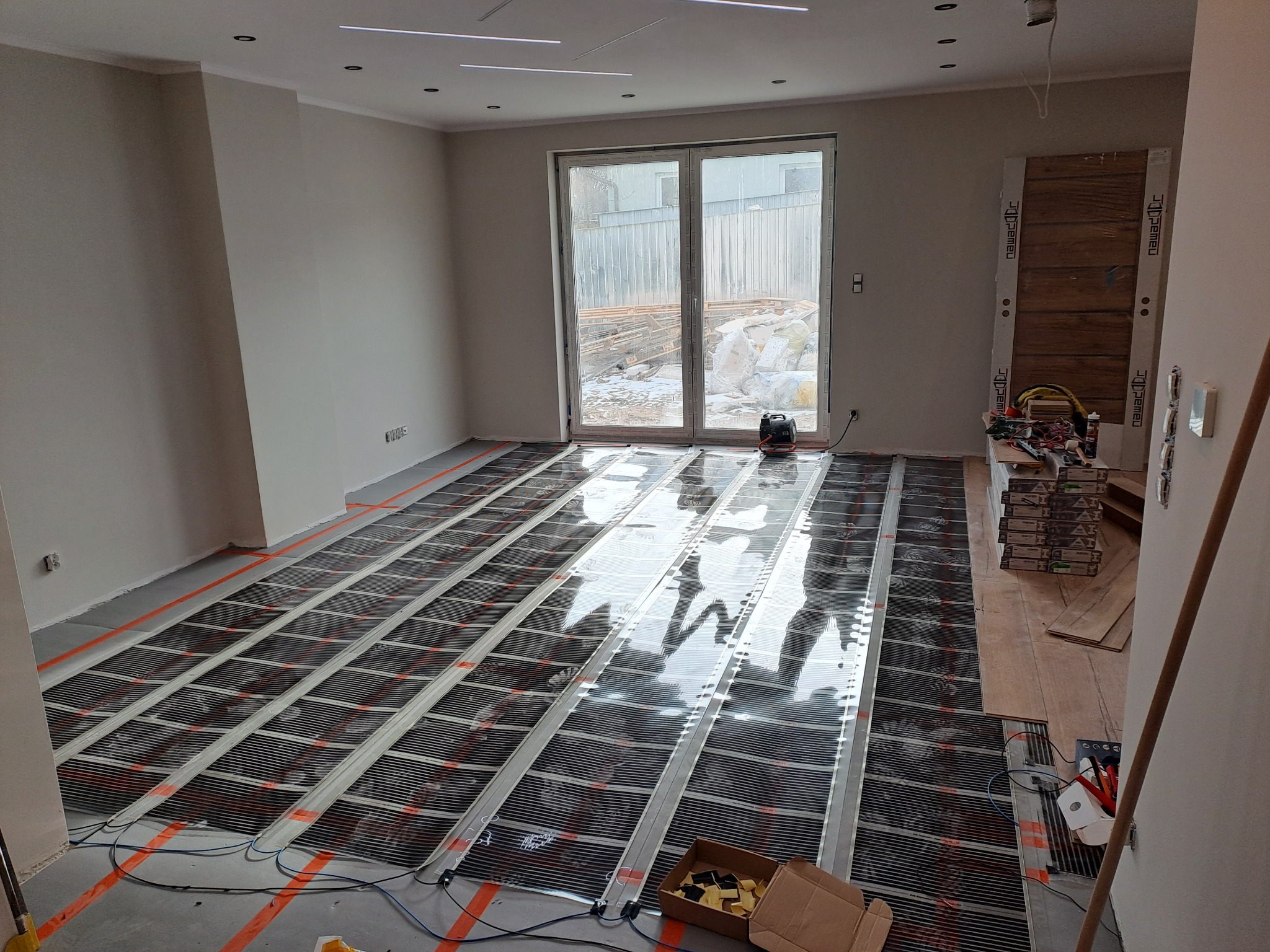 Przygotowanie do instalacji ogrzewania podłogowego: rozłożone maty grzewcze na podłożu w jasnym pokoju z dużym oknem balkonowym, widoczne narzędzia i materiały.