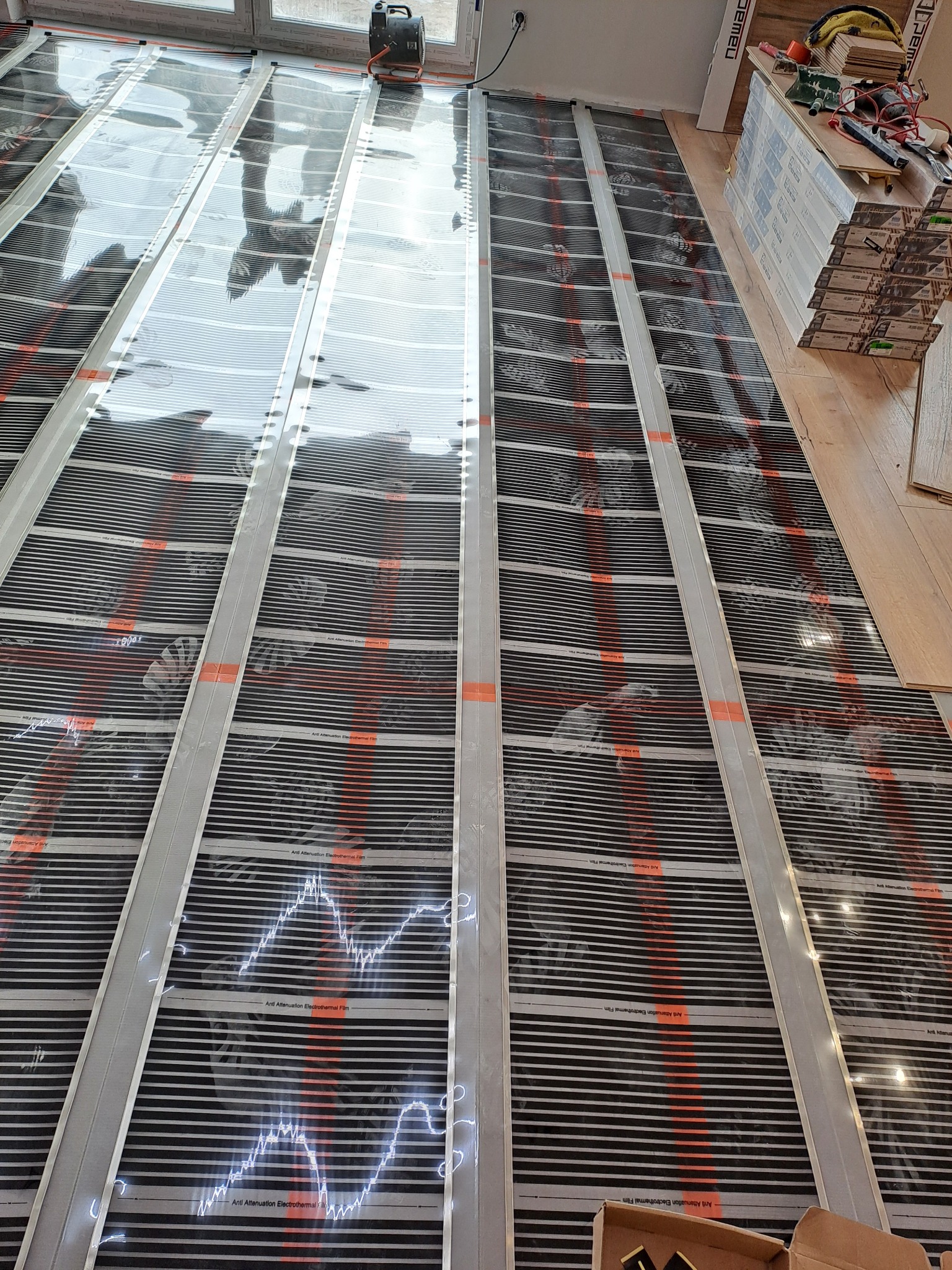 Montaż folii grzewczej pod podłogę, widoczne paski folii z elementami grzejnymi i napisy 'Anti Attenuation Electrothermal Film'. W tle narzędzia i materiały.