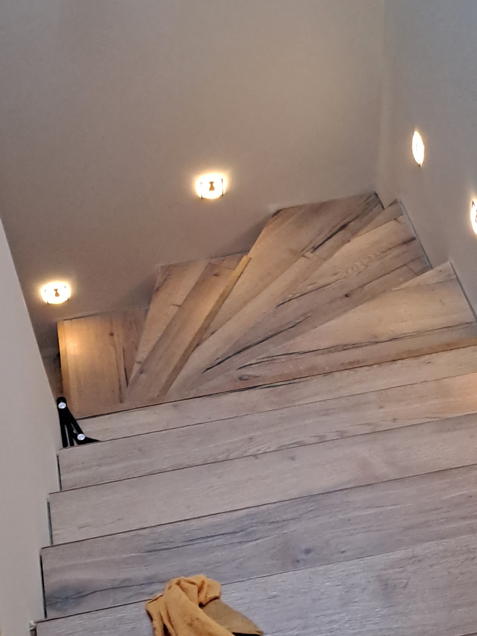 Drewniane schody spiralne z oświetleniem punktowym, wykończone panelami imitującymi drewno, widoczne narzędzia i ścierka. Układanie paneli na schodach.