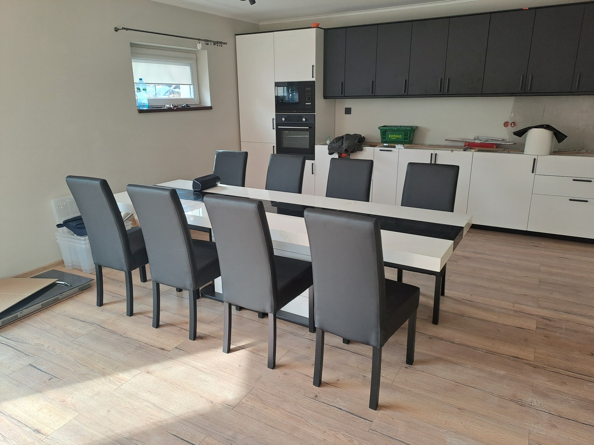 Jasne wnętrze po remoncie: stół z krzesłami na tle białych i grafitowych szafek kuchennych, drewnopodobna podłoga, okno z roletą.
