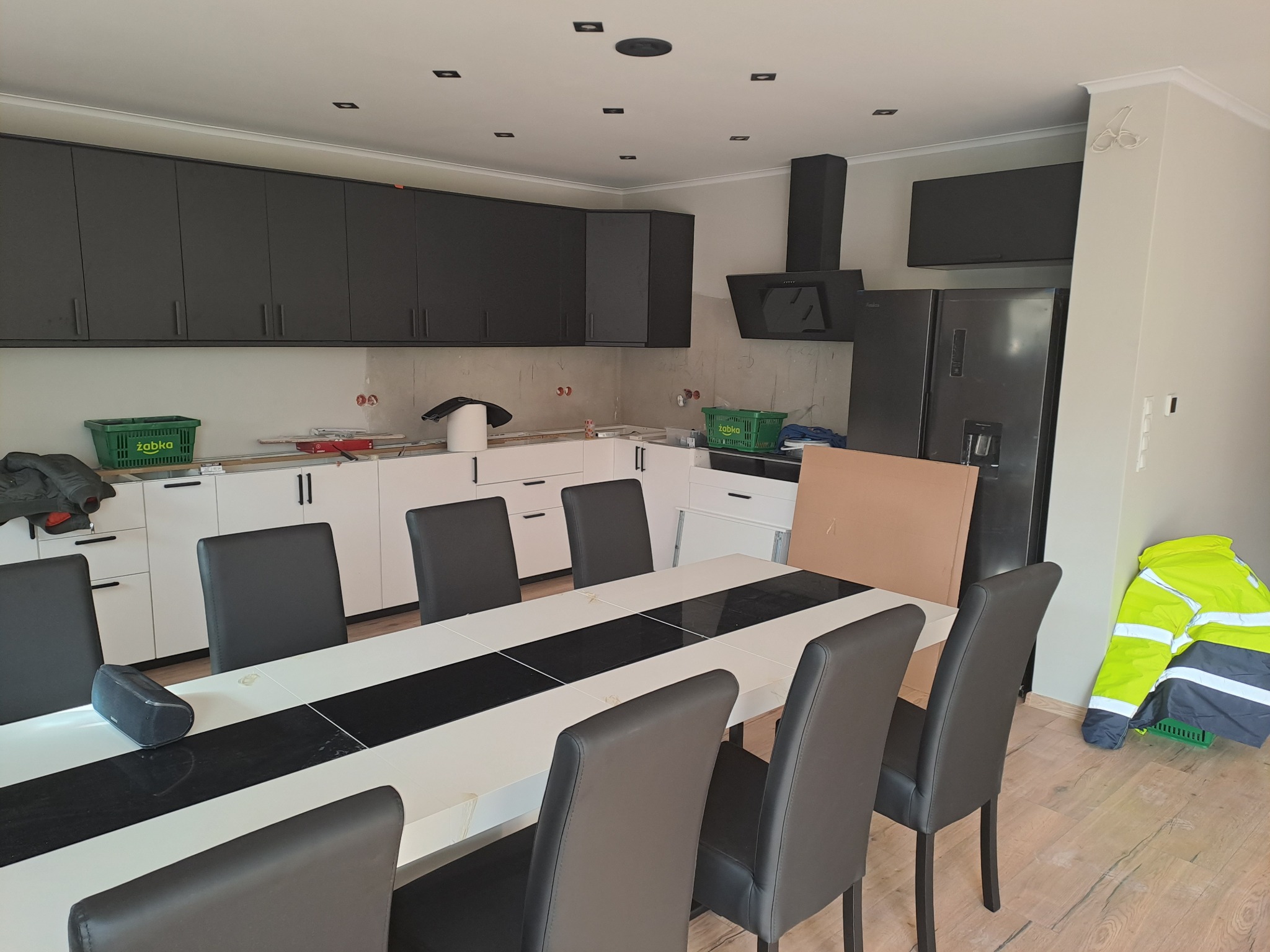 Wnętrze kuchni w trakcie wykończenia: białe szafki dolne, grafitowe górne, czarny okap i lodówka. Na pierwszym planie stół z krzesłami i odblaskowa kurtka.