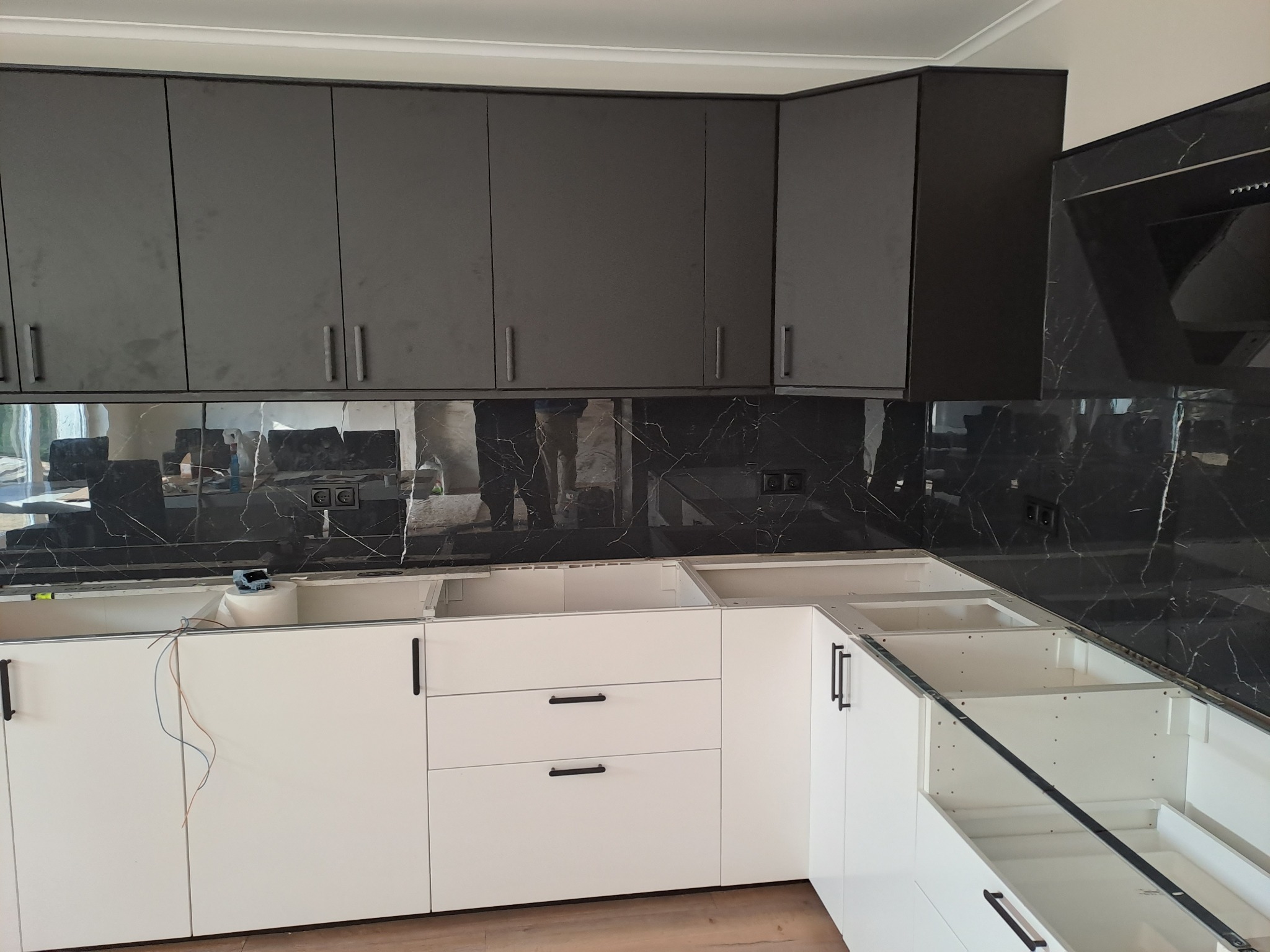 Nowoczesna kuchnia w trakcie montażu: szare szafki górne, białe dolne z czarnymi uchwytami, marmurowy panel ścienny, widoczne przewody elektryczne.