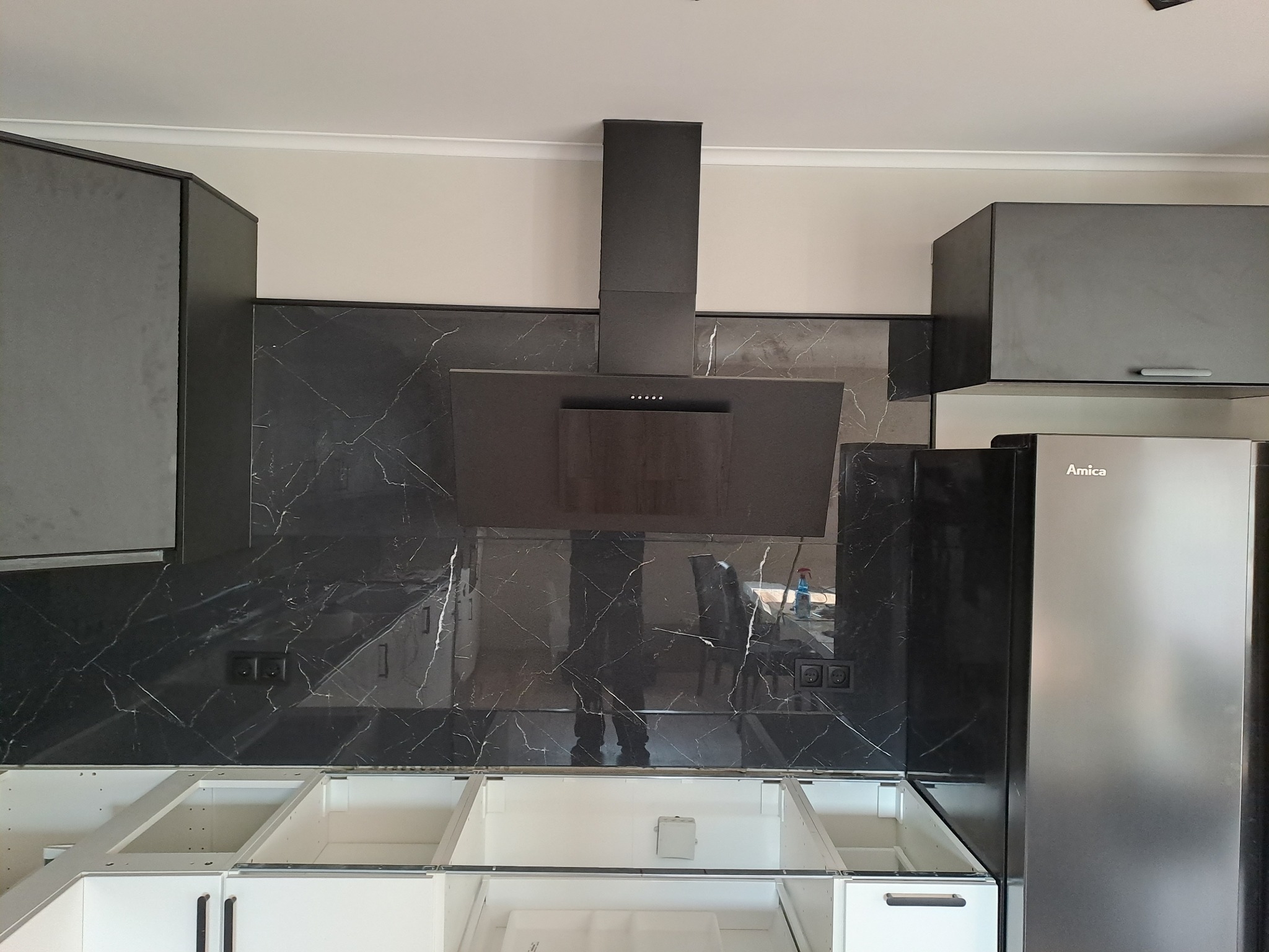 Częściowo zamontowana kuchnia z czarnym marmurowym panelem, szarymi szafkami i lodówką Amica. Widoczny montaż szafek dolnych i górnych oraz okap.
