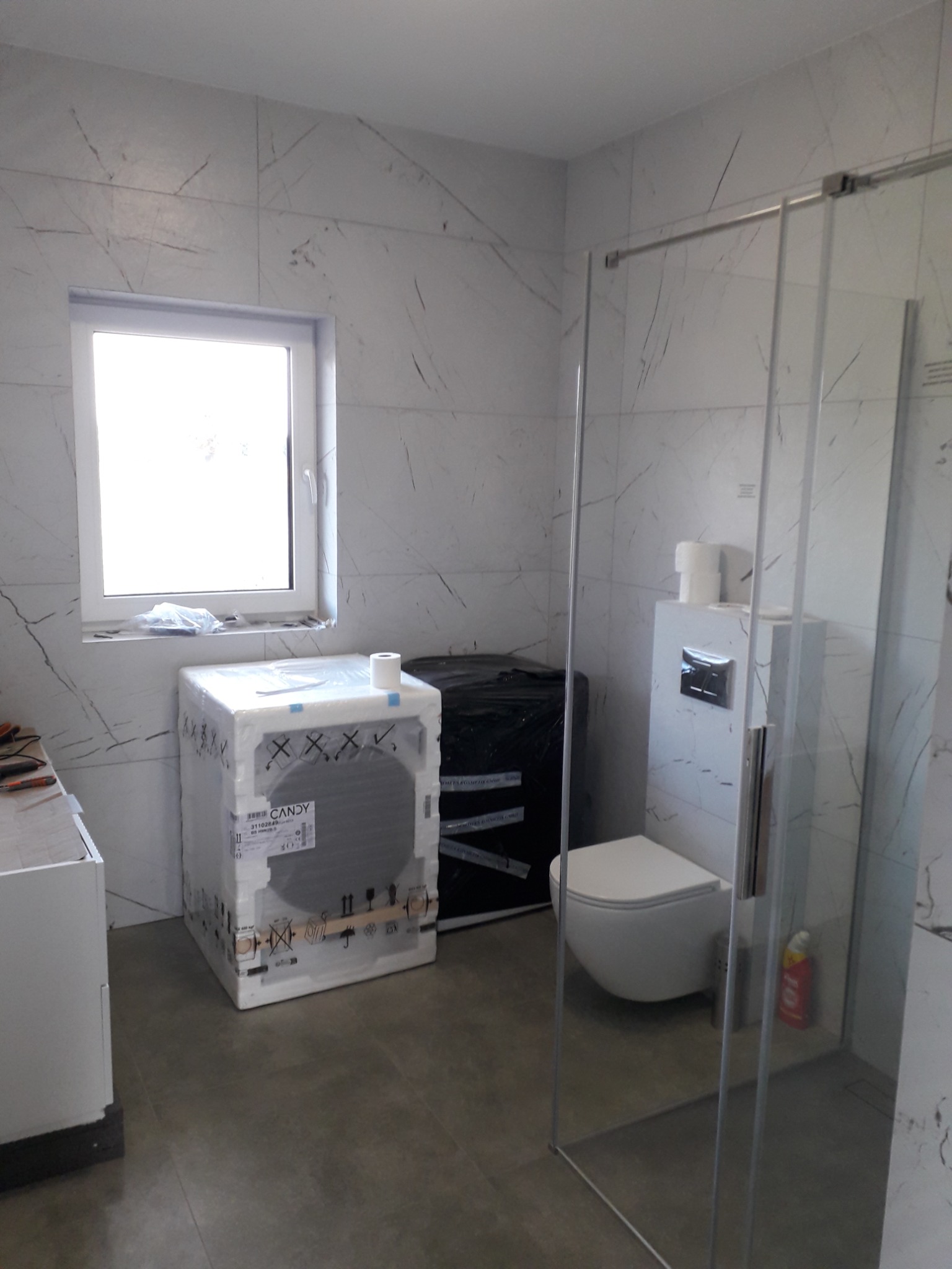 Nowa łazienka w trakcie wykańczania: kabina prysznicowa ze szkła, toaleta podwieszana, pralka Candy w opakowaniu i marmurowe płytki na ścianach.