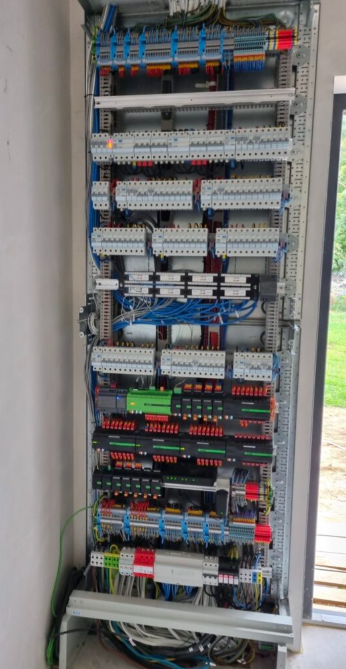Rozbudowana szafa elektryczna z licznymi zabezpieczeniami, przewodami i modułami, zamontowana w nowym budynku. Widoczne elementy automatyki i okablowanie.