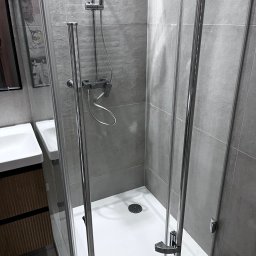 Usługi remontowo budowlane Roman Kapituła - Nowoczesna kabina prysznicowa z szarymi płytkami, chromowanymi detalami i białym brodzikiem. Widoczna umywalka z szafką. Minimalistyczny design, czyste linie, świeżo po remoncie.