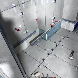 Usługi remontowo budowlane Roman Kapituła - Jasnoszare płytki w trakcie układania z użyciem systemu poziomowania. Widoczne narzędzia: młotek, poziomica, kielnia. Fragment zabudowy z profili.